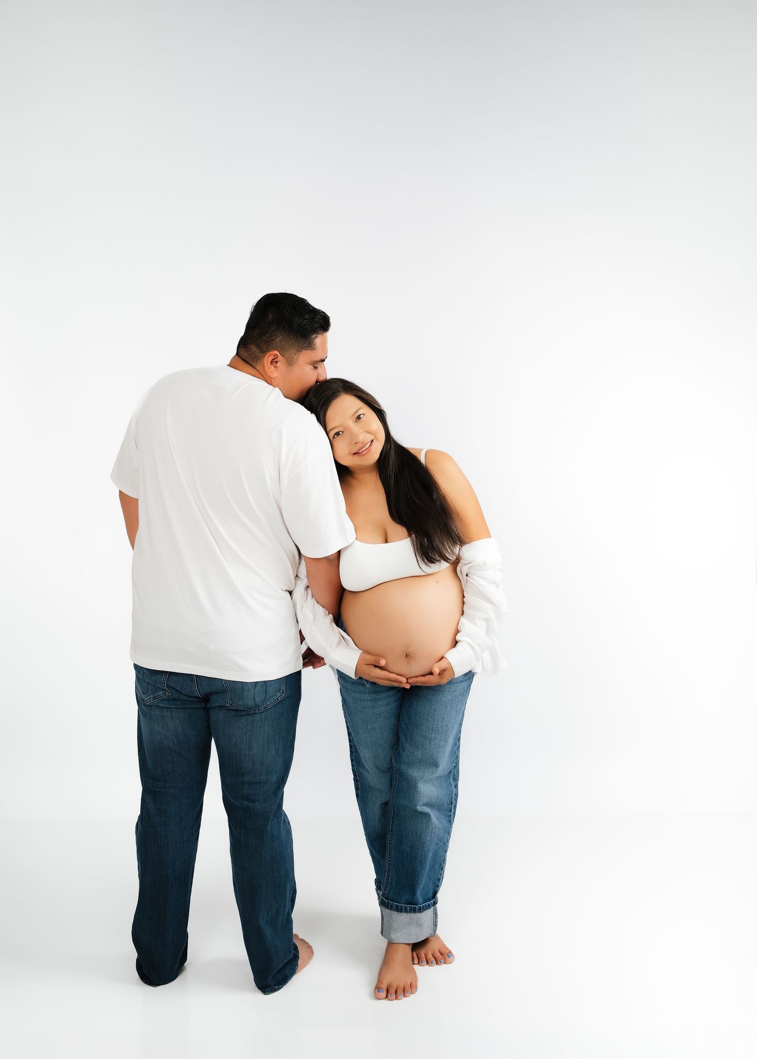 A pregnant couple in white tops and jeans pose tenderly, man kissing woman's head as she cradles her baby bump.