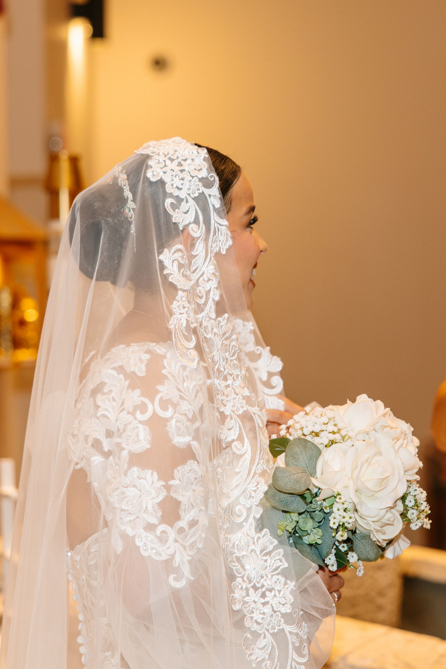 Bride in lace wedding gown and veil holds white rose bouquet at church ceremony.