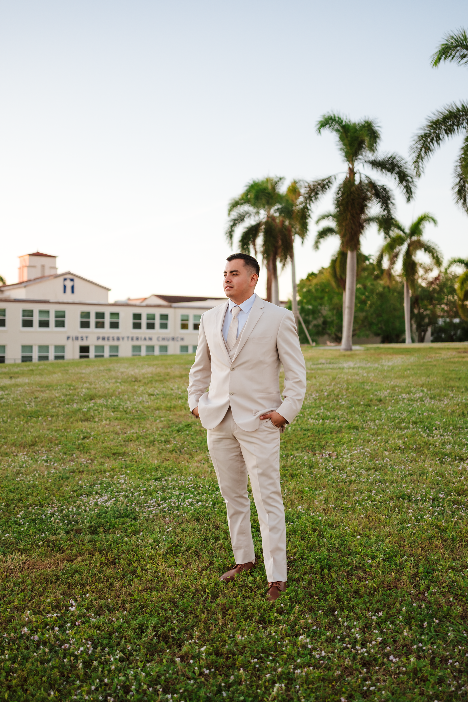 Handsome groom in beige suit standing on green lawn with palm trees and First Presbyterian Church in background.