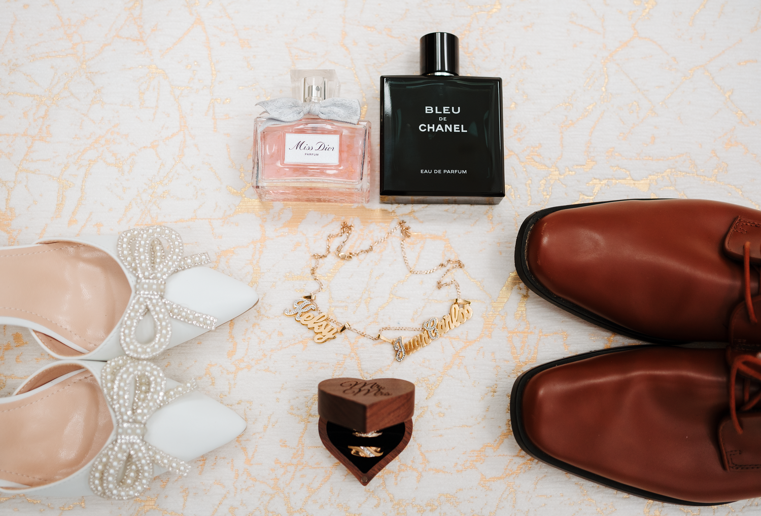 Flat lay of wedding accessories including Chanel perfume, gold bracelet, brown dress shoes, and heels.