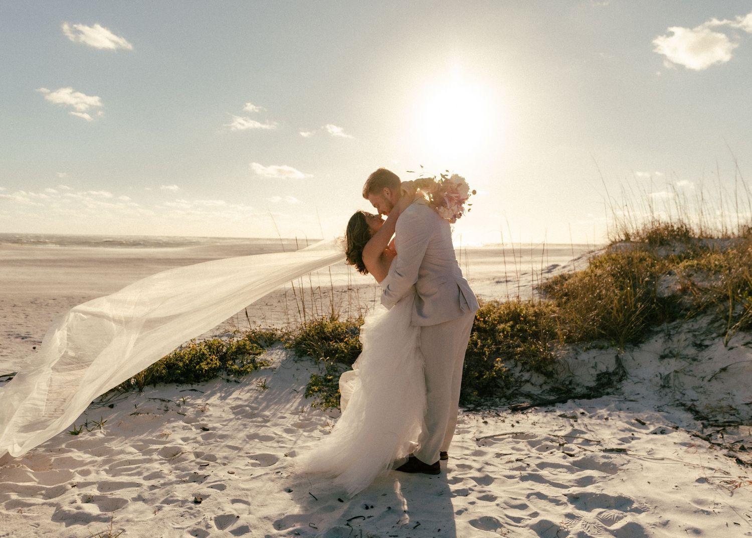 Bride and groom share a romantic kiss on a sunny beach, with veil flowing in the wind against a golden sky.