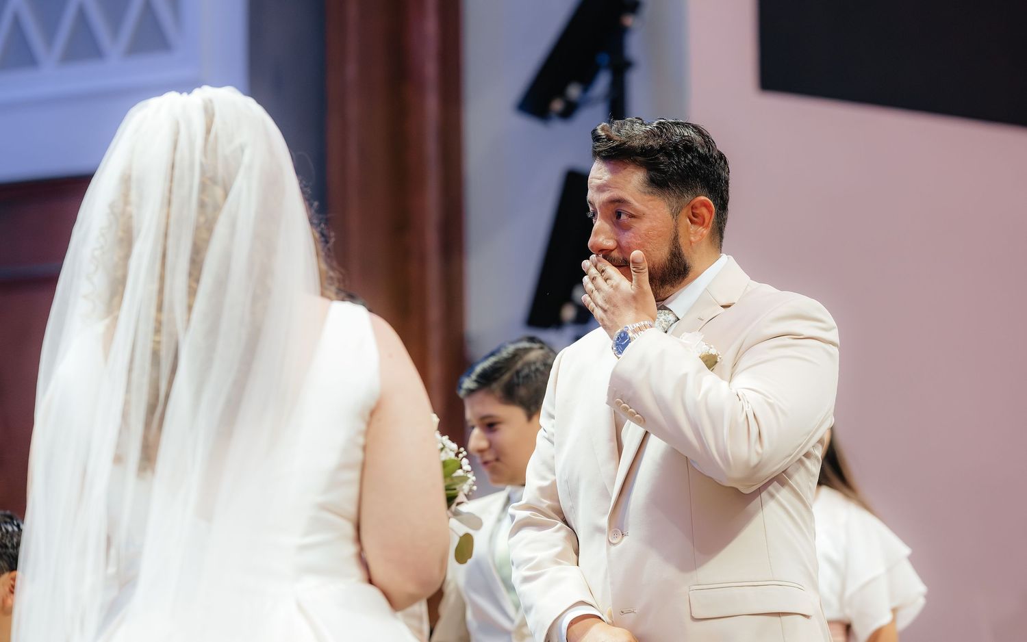 Emotional groom in beige suit kisses his hand while seeing bride in white wedding dress and veil at church ceremony.