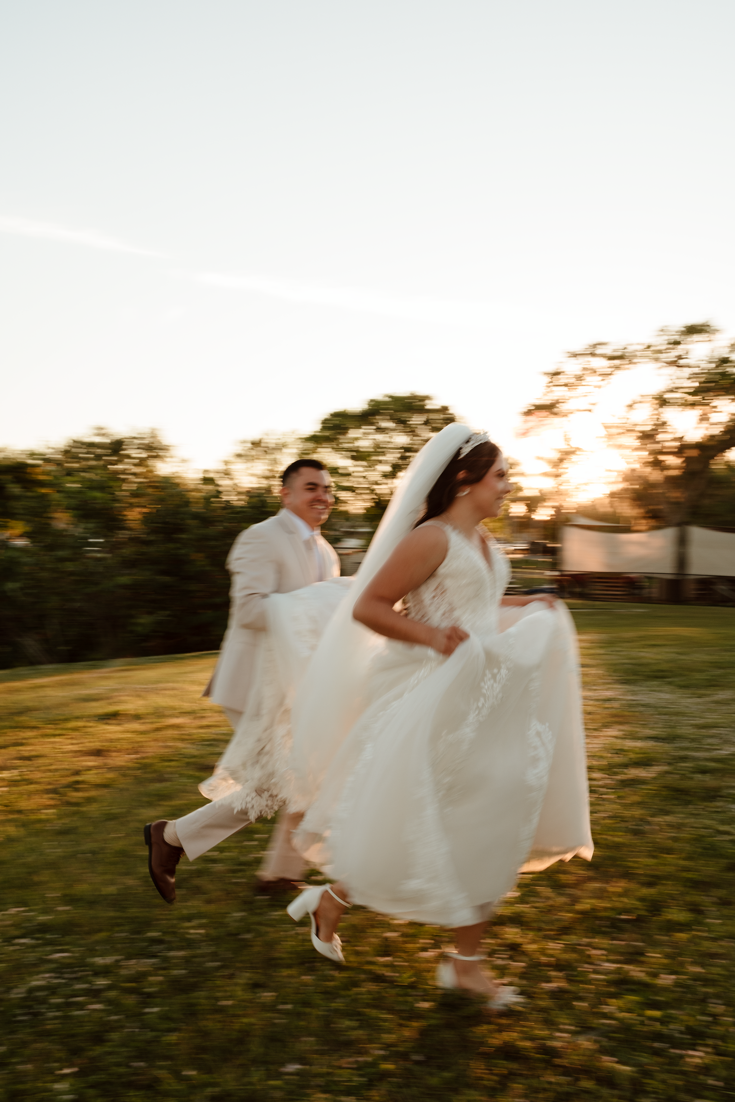 Bride and groom joyfully running through a grassy field at golden hour sunset.