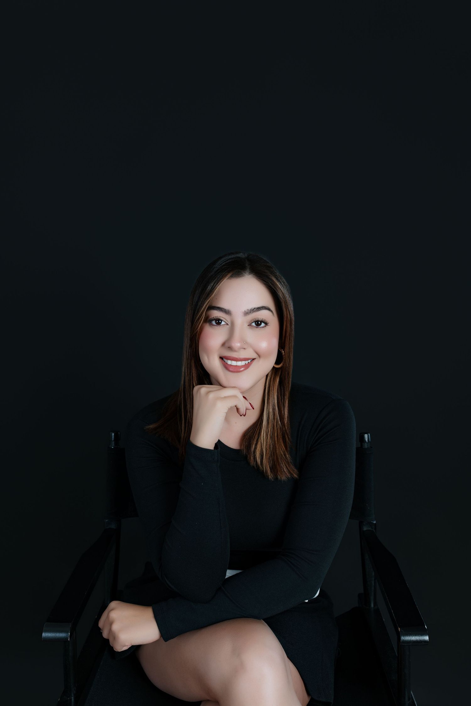 Young woman in black dress sitting on chair smiling with hand on chin against dark background.