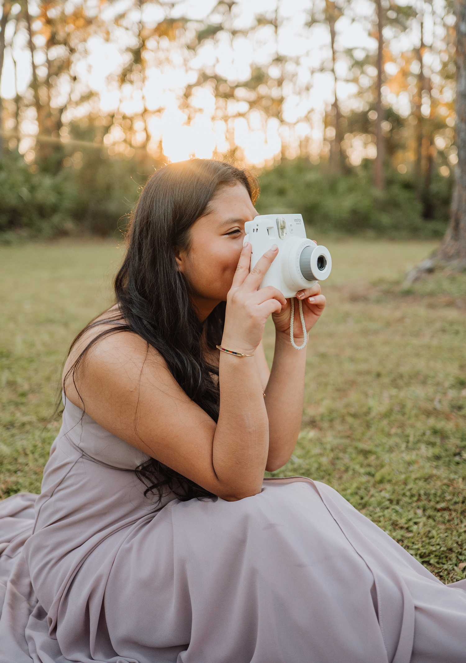 Woman in a lavender dress sits on the grass holding a white instant camera to her face at golden hour in a sunlit park.