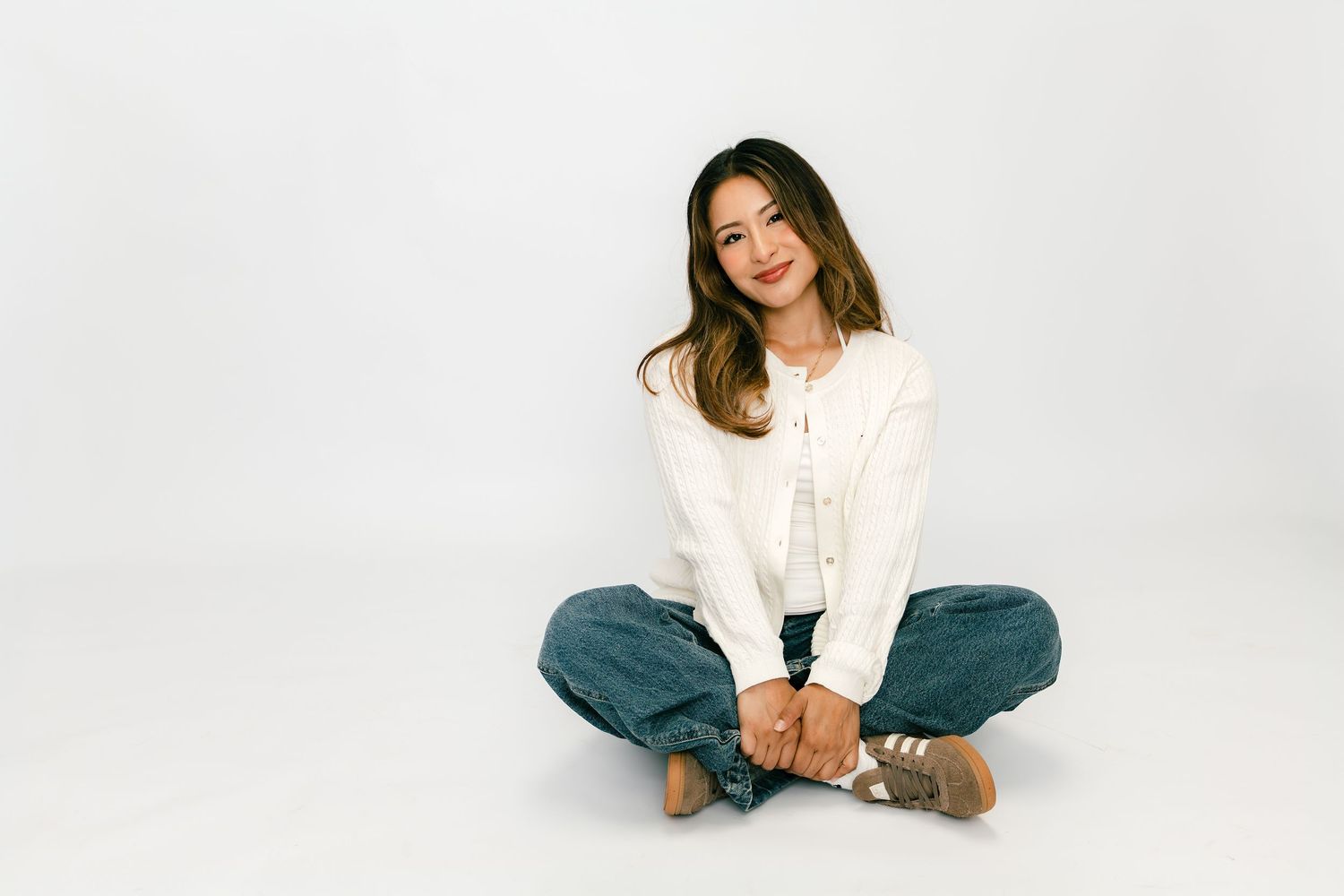 Young woman sitting cross-legged on white background, wearing white cardigan and blue jeans, smiling at camera.