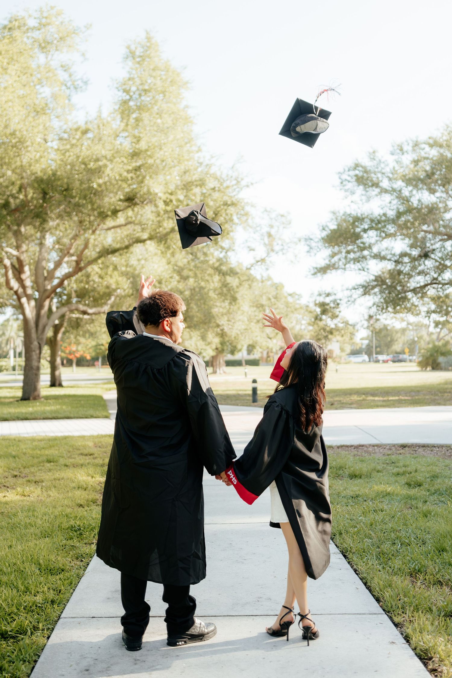 Two graduates in black gowns toss their caps in the air on a sunny campus walkway.