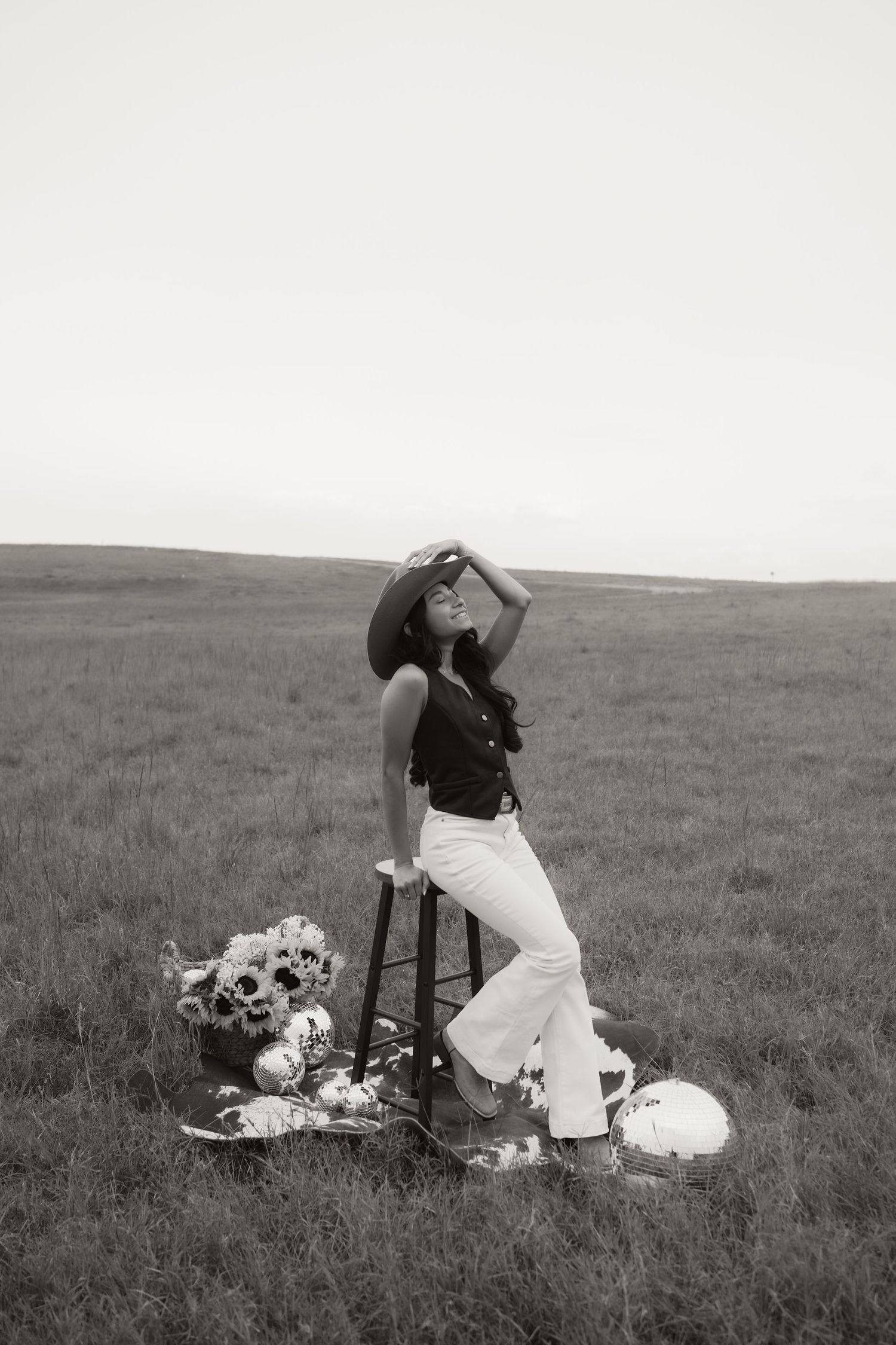Black and white photo of a woman in wide-leg pants and cowboy hat posing on a stool in an open field with flowers.