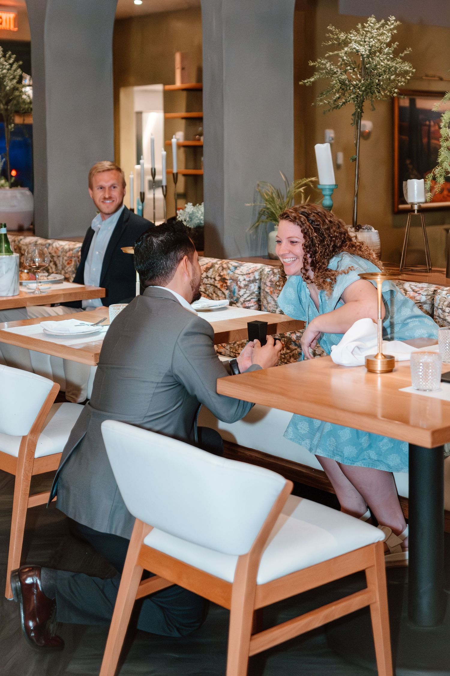 Man proposes to woman at elegant restaurant table while smiling waiter watches in the background.