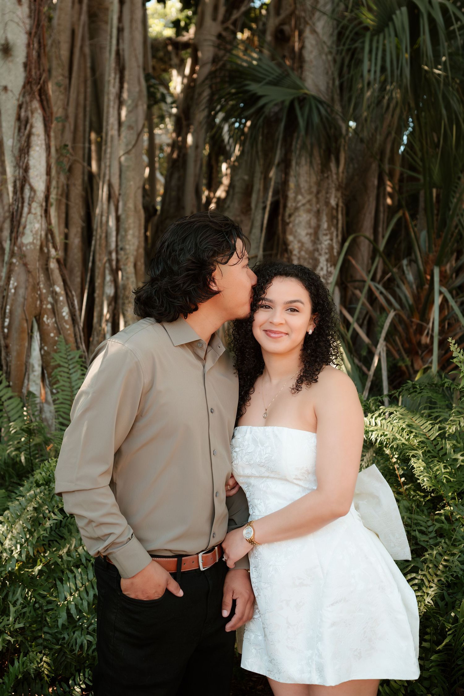 A couple poses romantically in a lush tropical garden, man kissing woman's cheek as she smiles in a white dress.