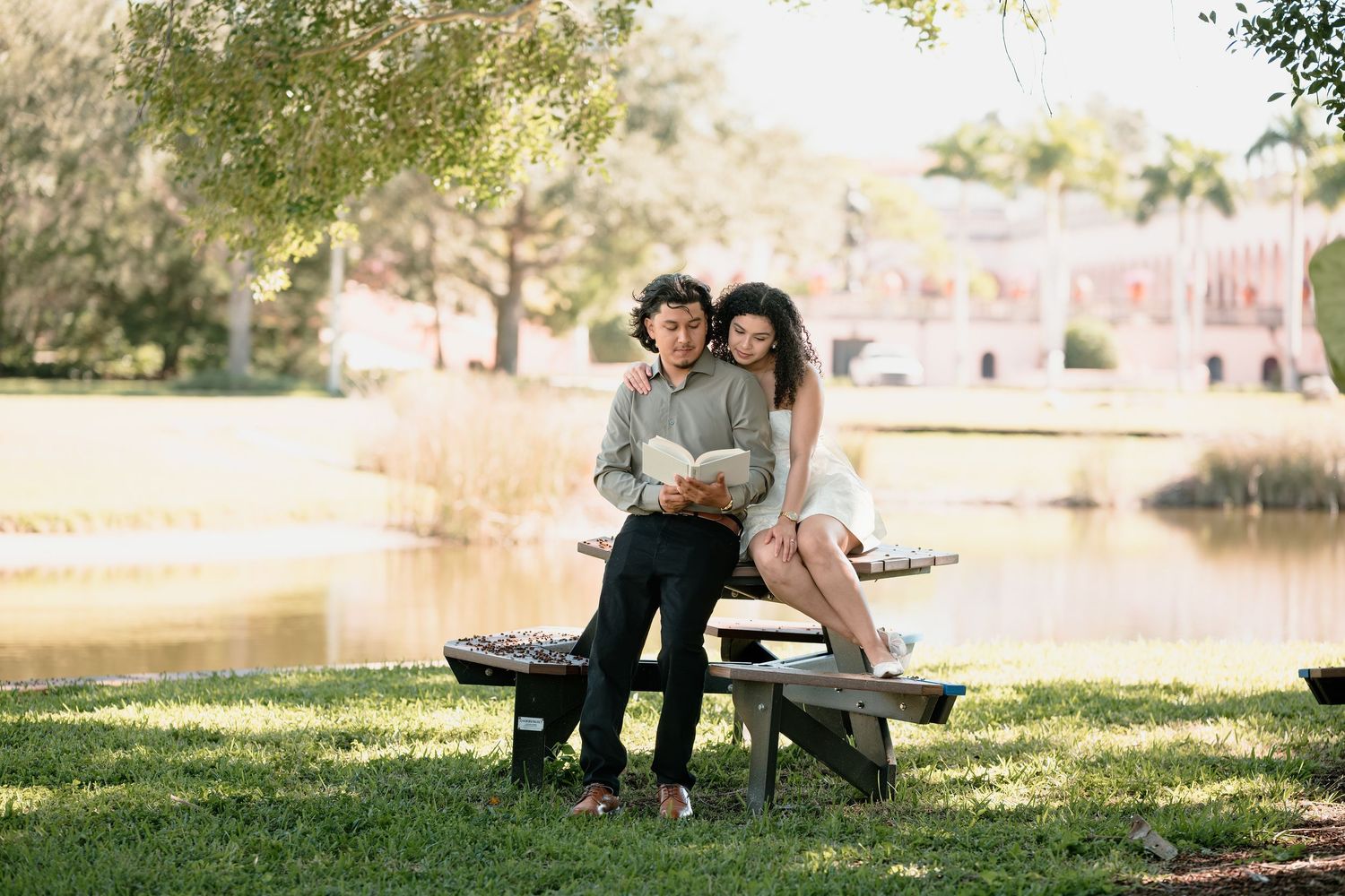 Couple sitting on a picnic bench by a lake reading a book together in a lush green park near a pink building.