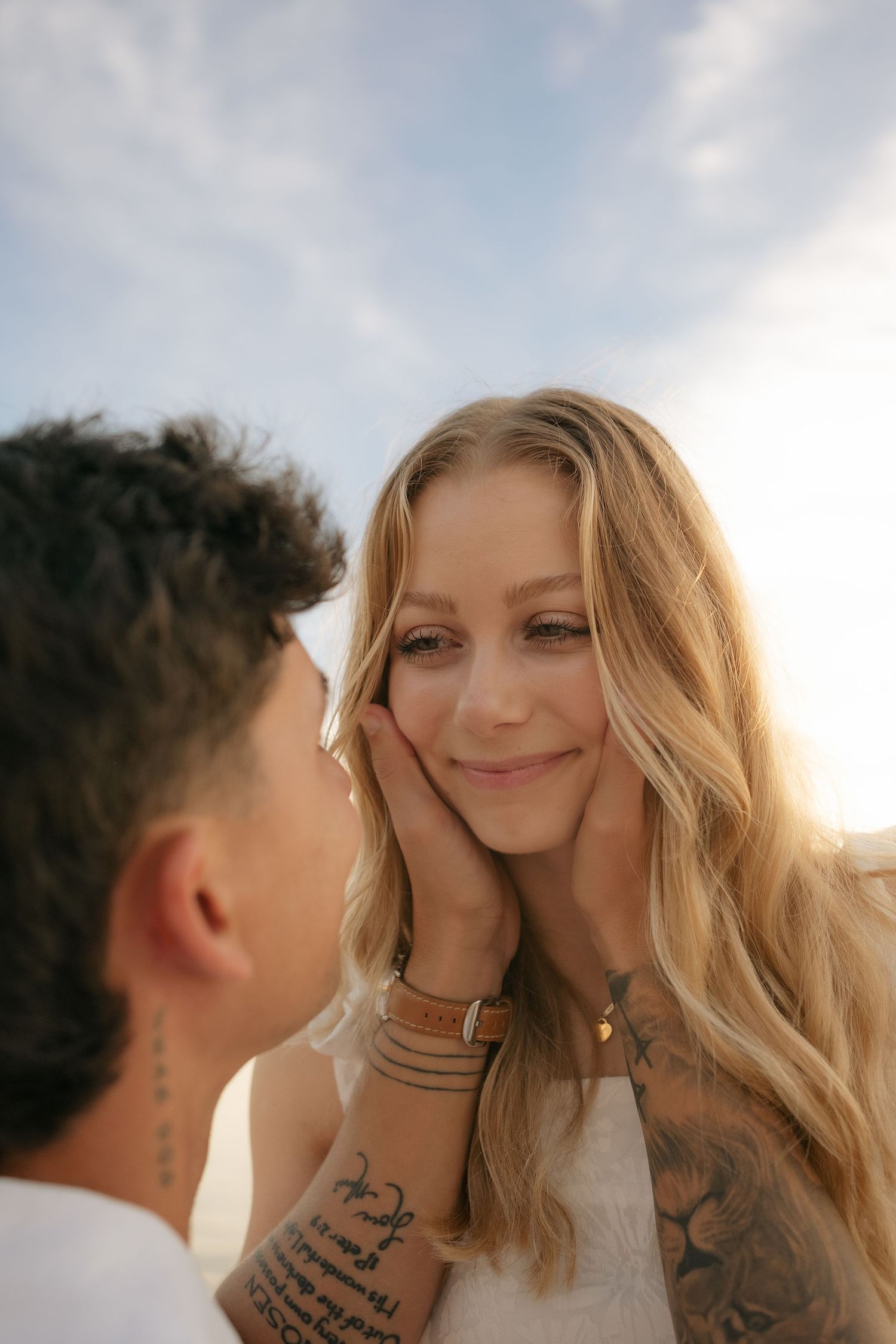 A tattooed couple shares a tender moment outdoors, man gently cupping womans face, backlit by golden sunlight.