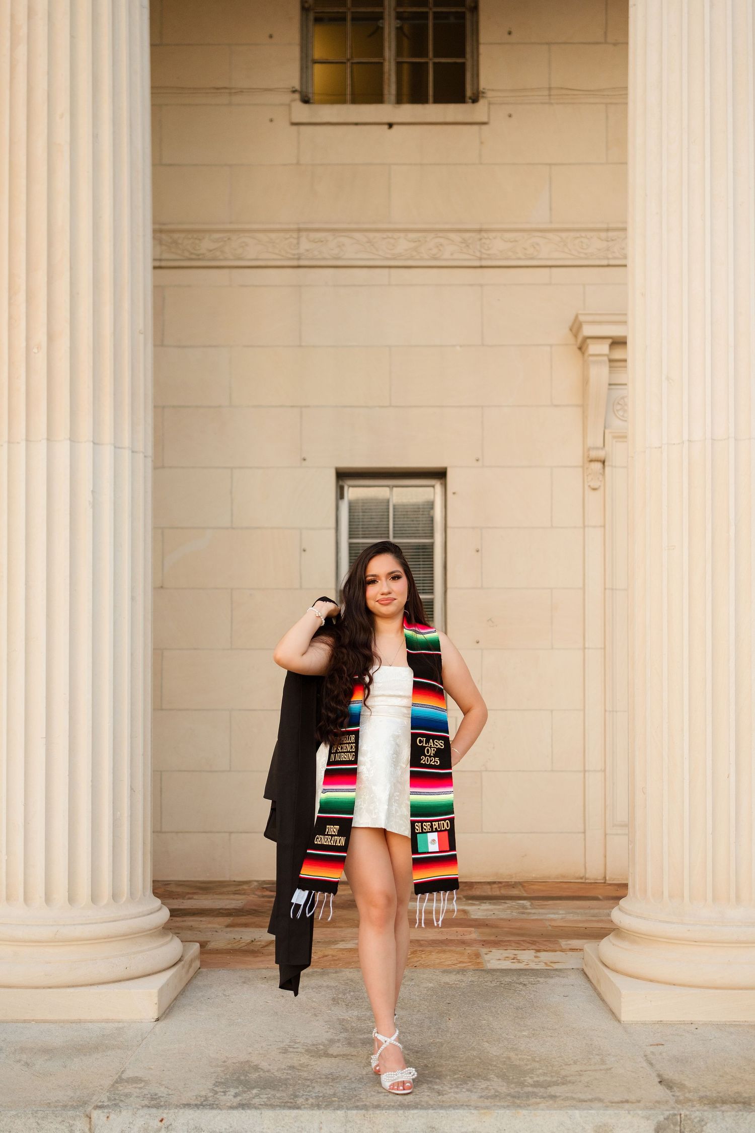 Young woman in white dress with colorful graduation stole and cap poses between columns for senior portrait.