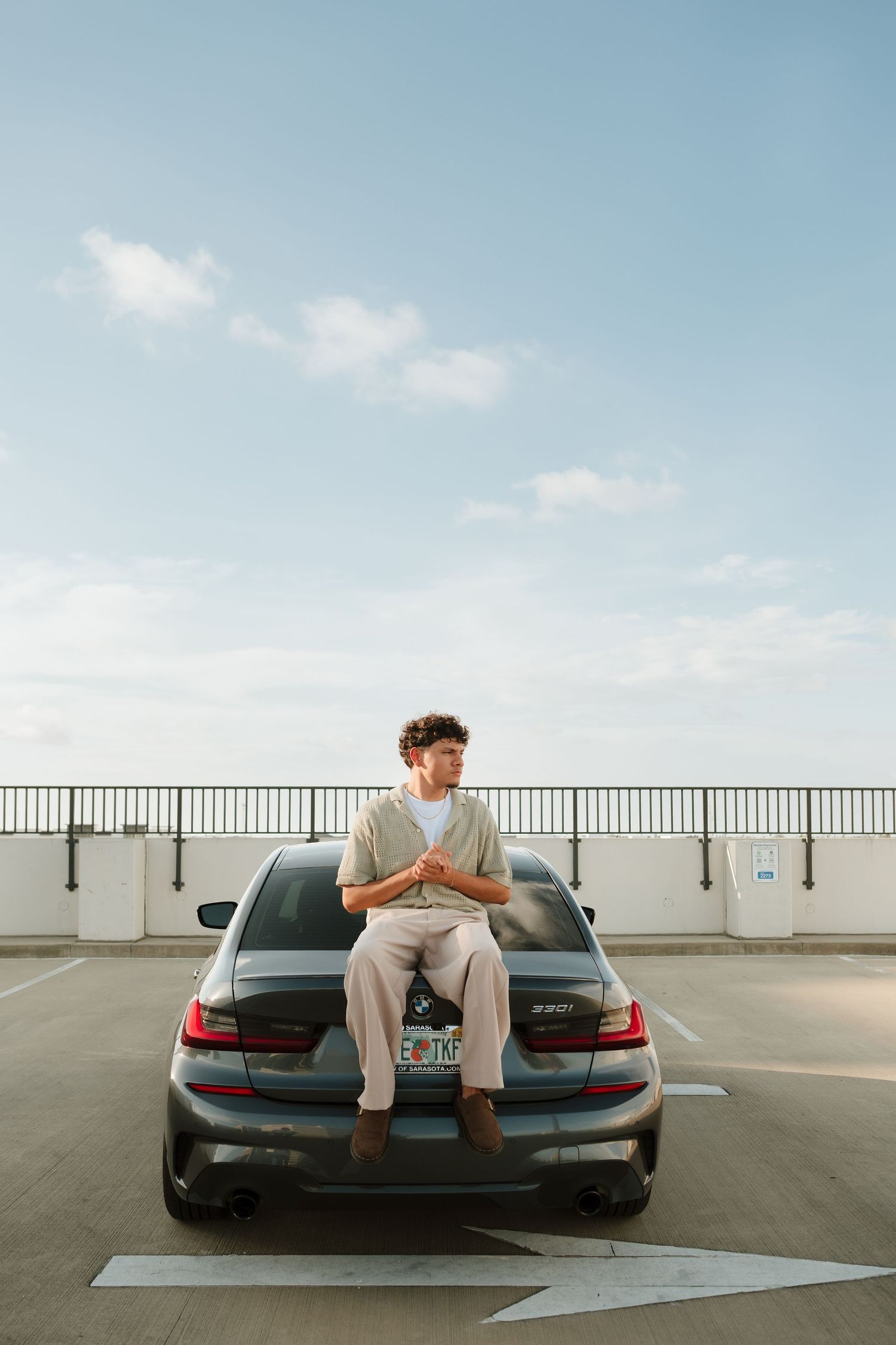 Young man with curly hair sits on trunk of gray BMW sedan in rooftop parking lot under blue sky.