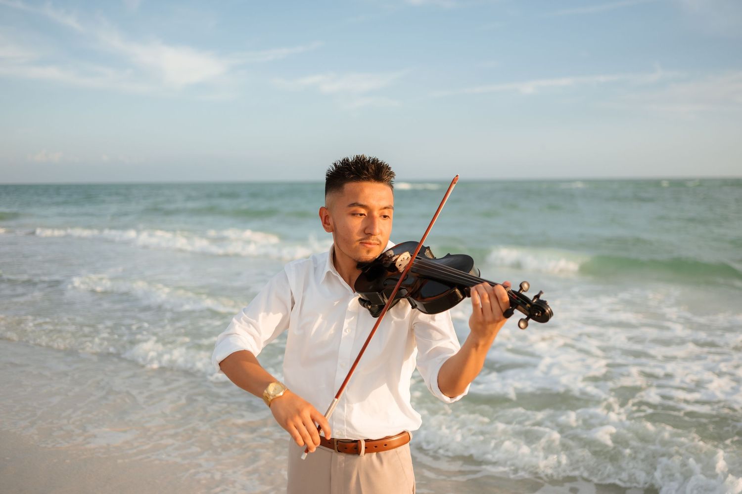 Young Asian man playing violin on a sunny beach with ocean waves, wearing a white shirt and khakis.