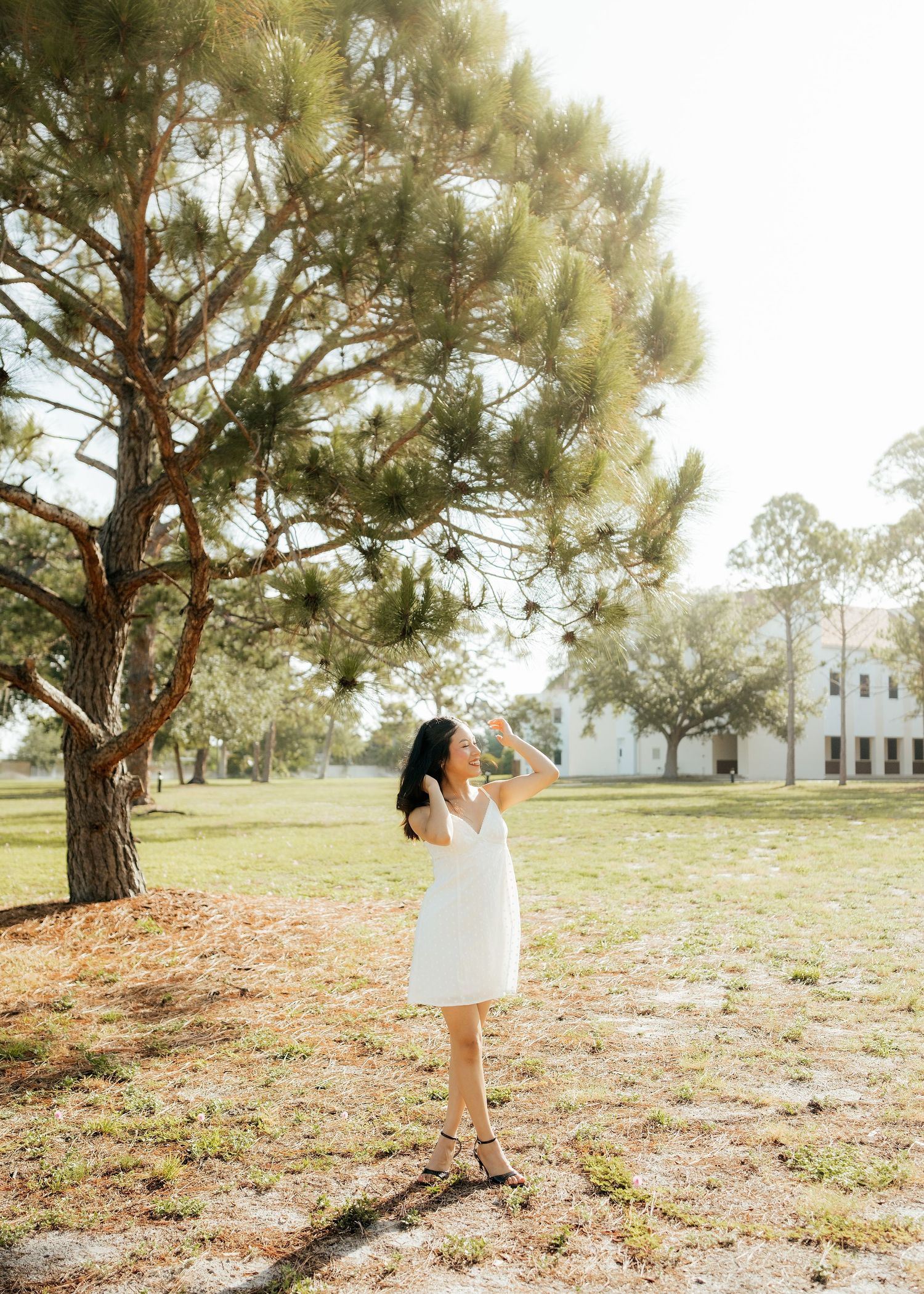 Young woman in white dress standing under pine tree in sunlit park, looking up with arms raised.