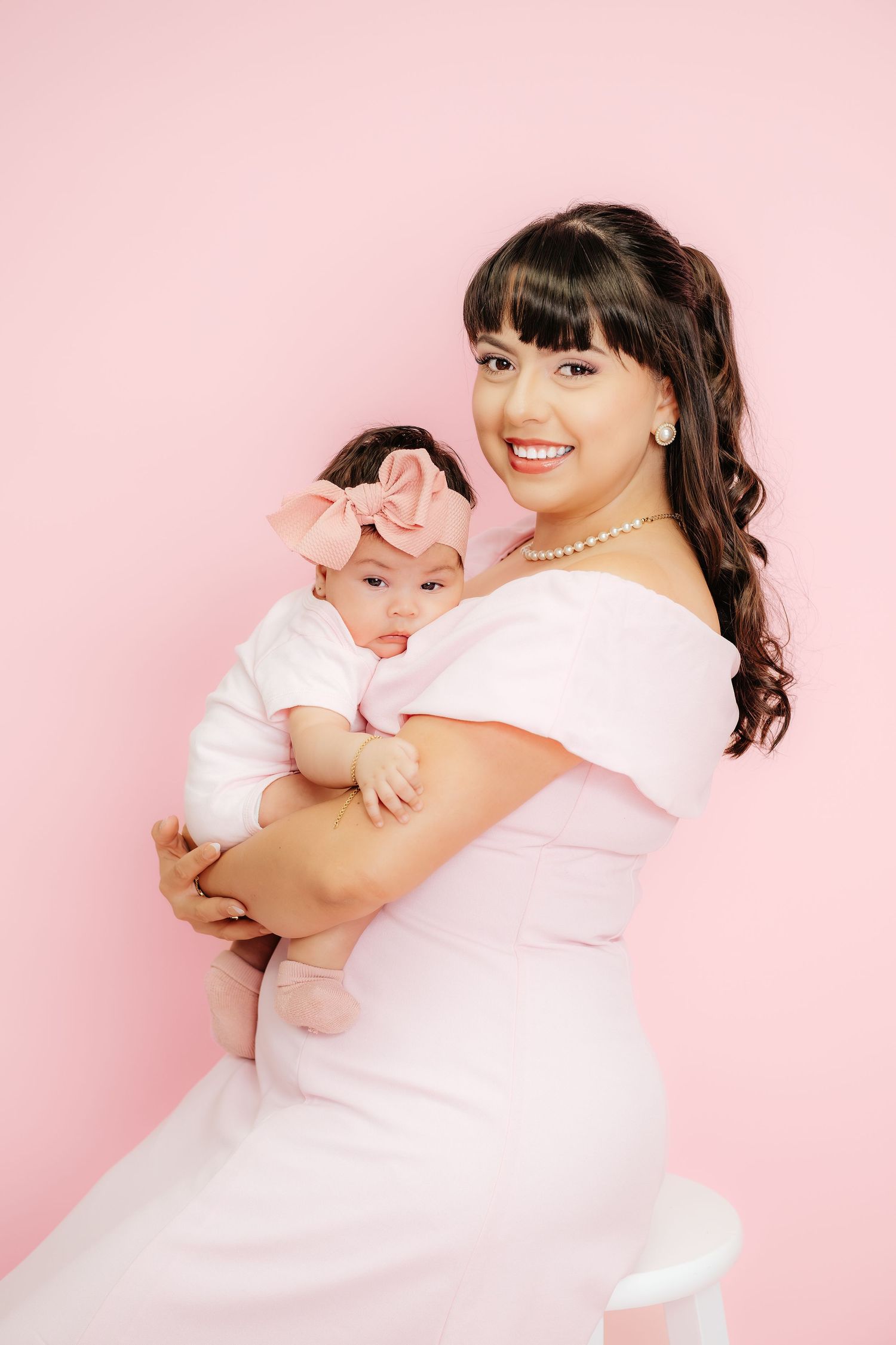 A smiling woman in a pink off-shoulder dress holds her baby girl wearing a pink bow headband against a pink background.