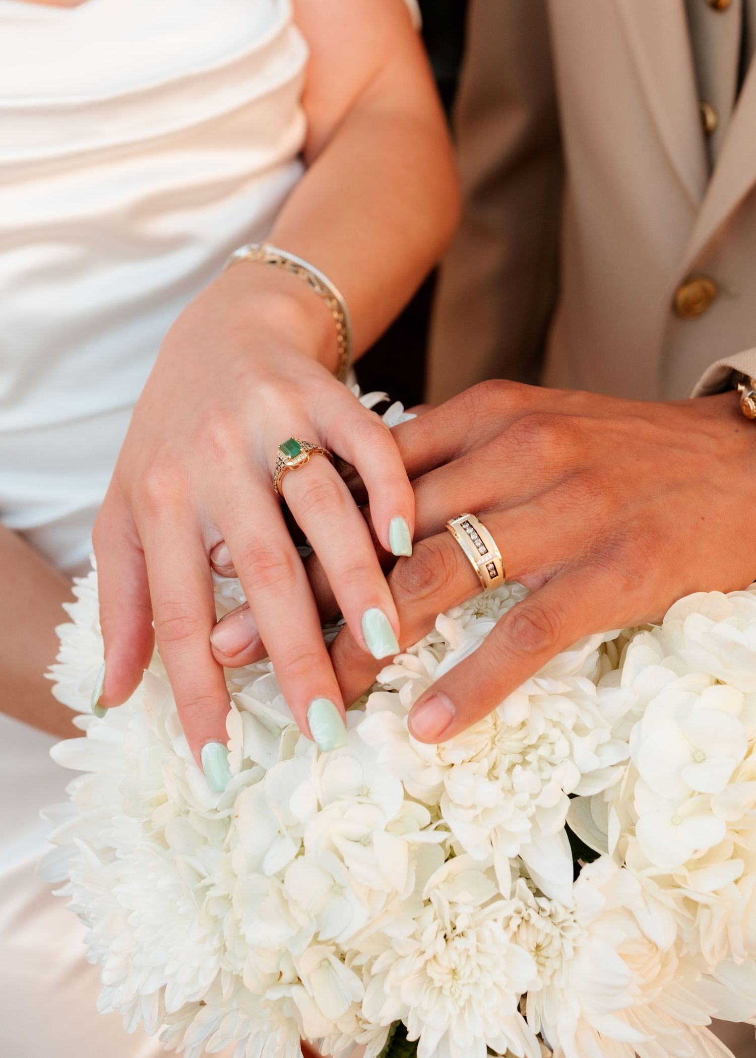 Bride and groom displaying wedding rings on white floral bouquet, featuring emerald engagement ring and gold band.