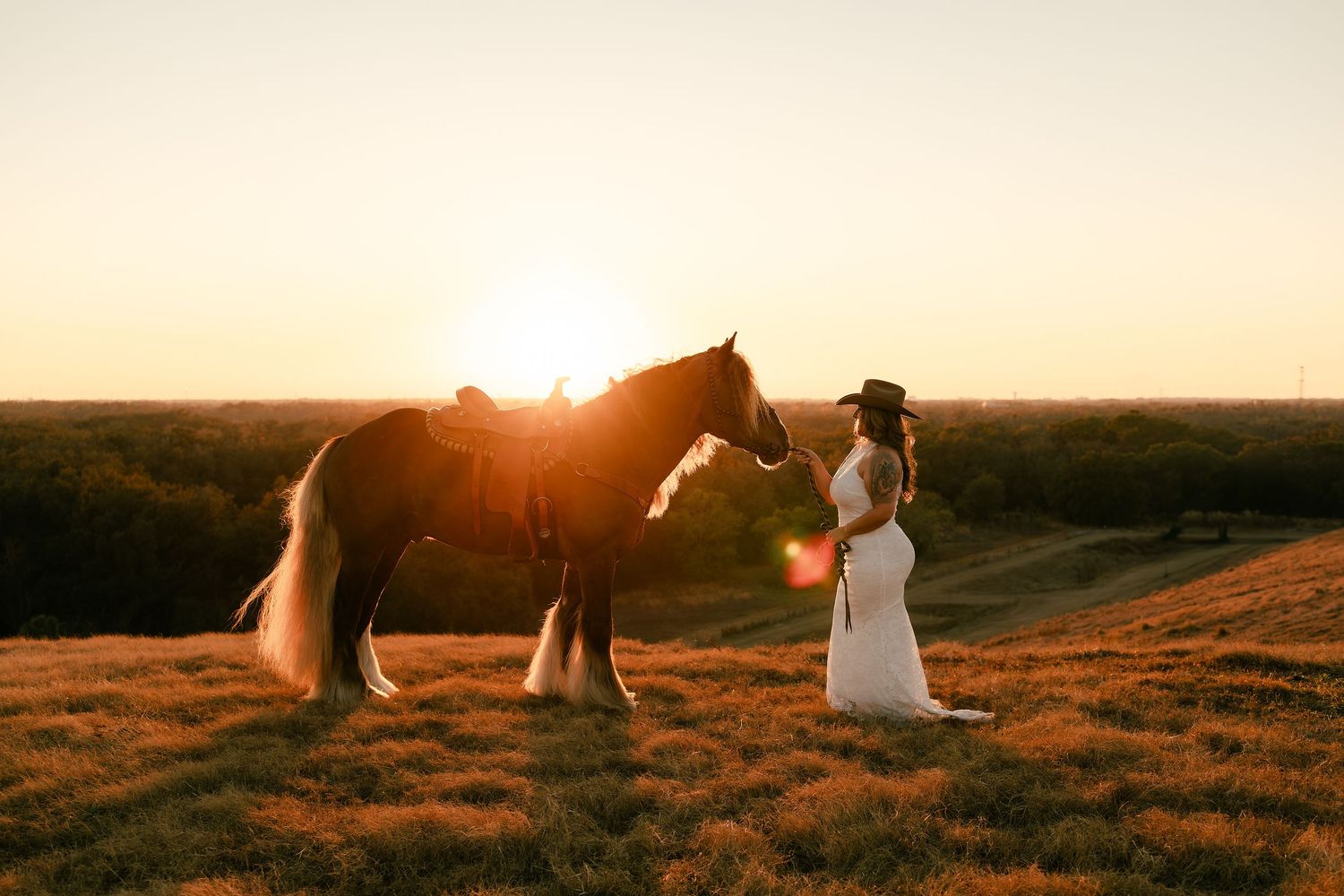 Woman in white dress and cowboy hat stands with a brown horse at golden sunset on a grassy hill.