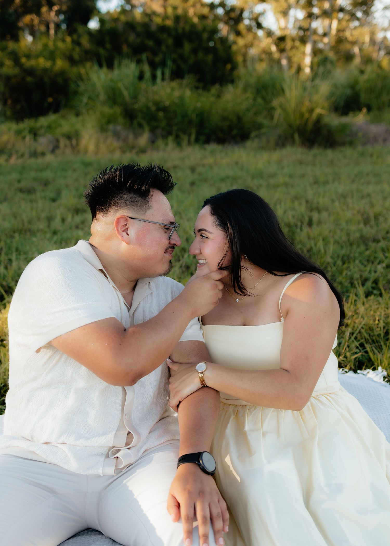 Couple in white outfits sitting on grass, sharing a tender moment outdoors during golden hour.