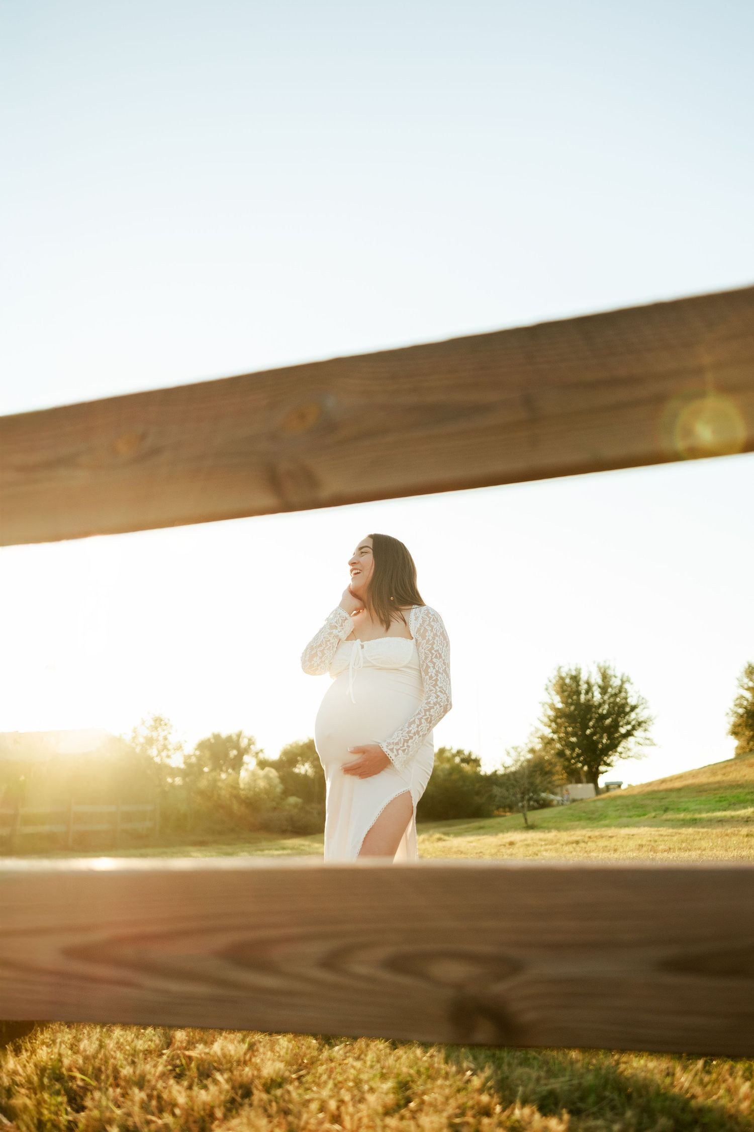 A woman in a white dress stands behind a wooden fence at sunset with golden light illuminating a rural field.