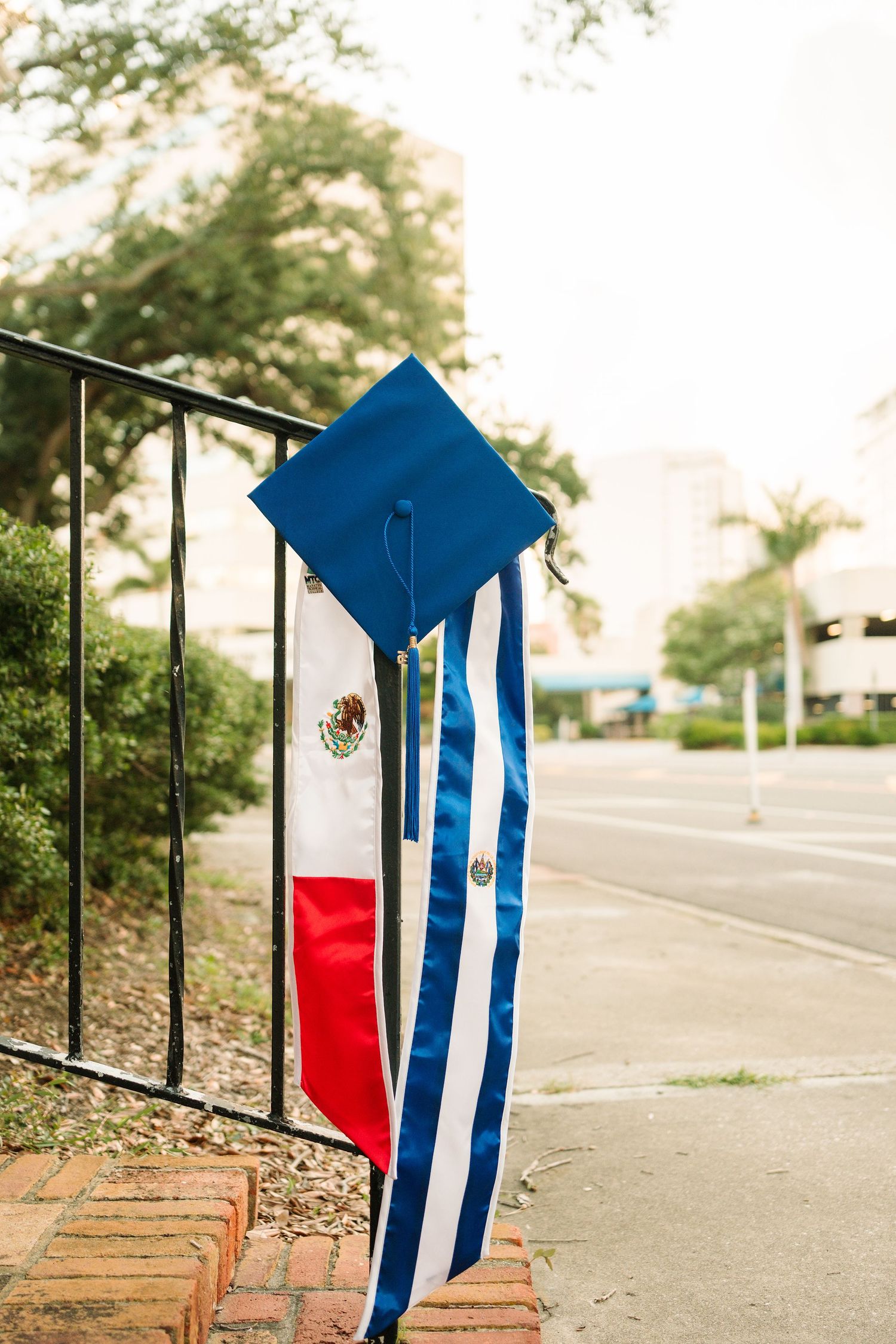A blue graduation cap rests against a metal railing with a Mexican flag in the background.