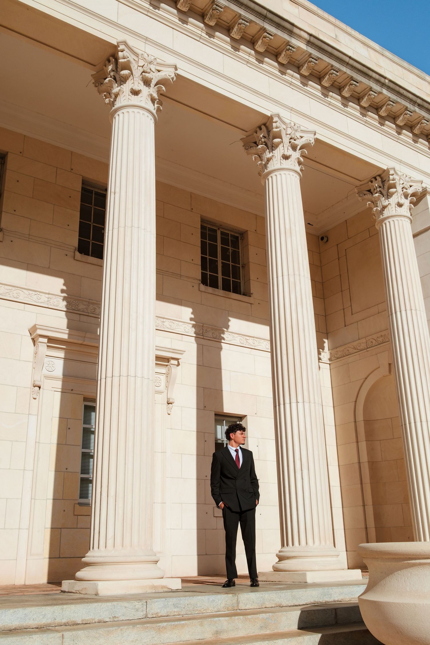 A young man in a black suit stands on steps between tall Corinthian columns of a neoclassical building.