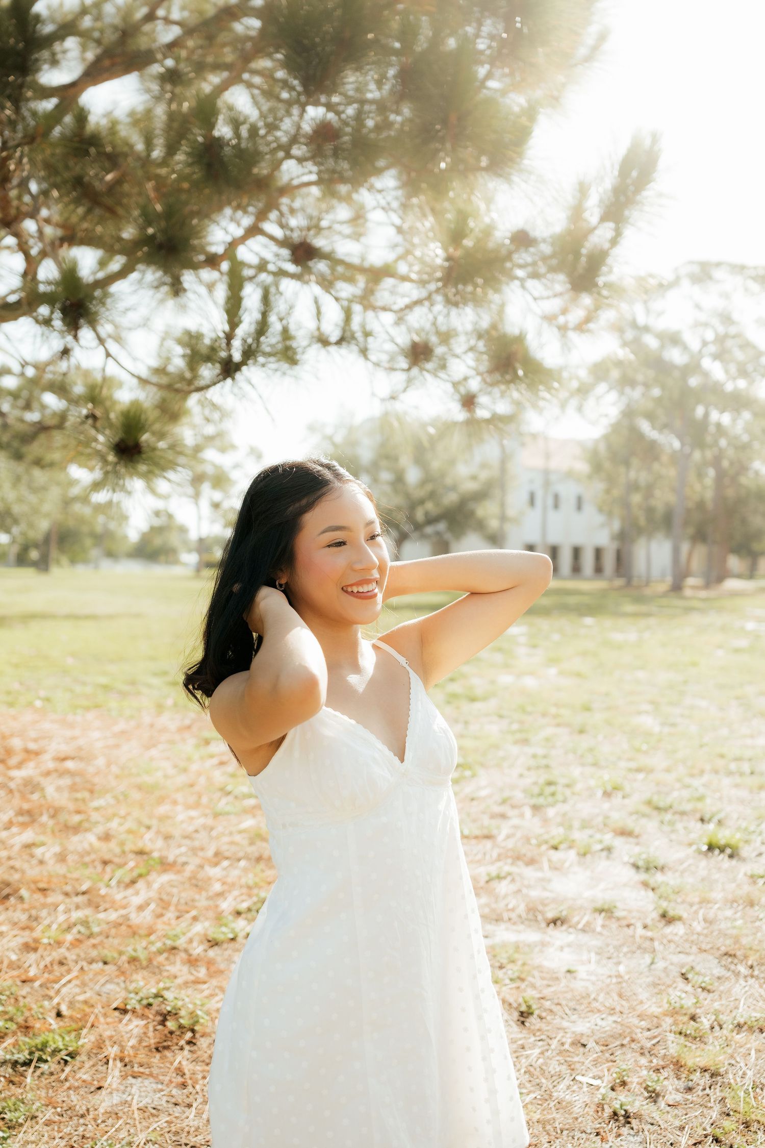 Smiling young woman in white sundress poses outdoors in sunlit park with trees and historic building in background.