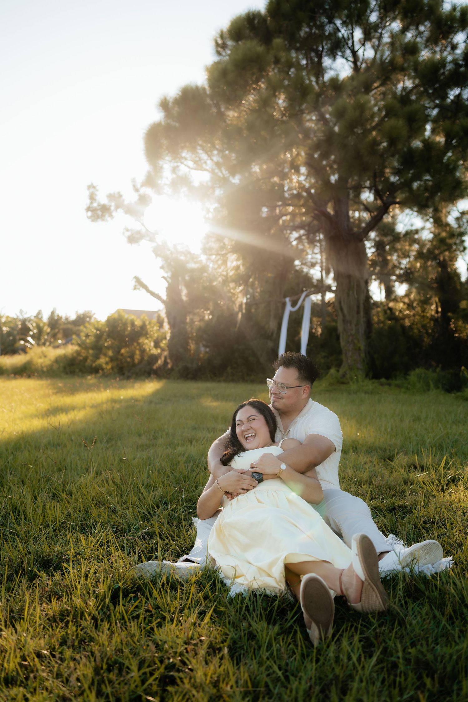 A happy couple sits together on green grass at golden hour, laughing with a pine tree and soft sunlight behind them.