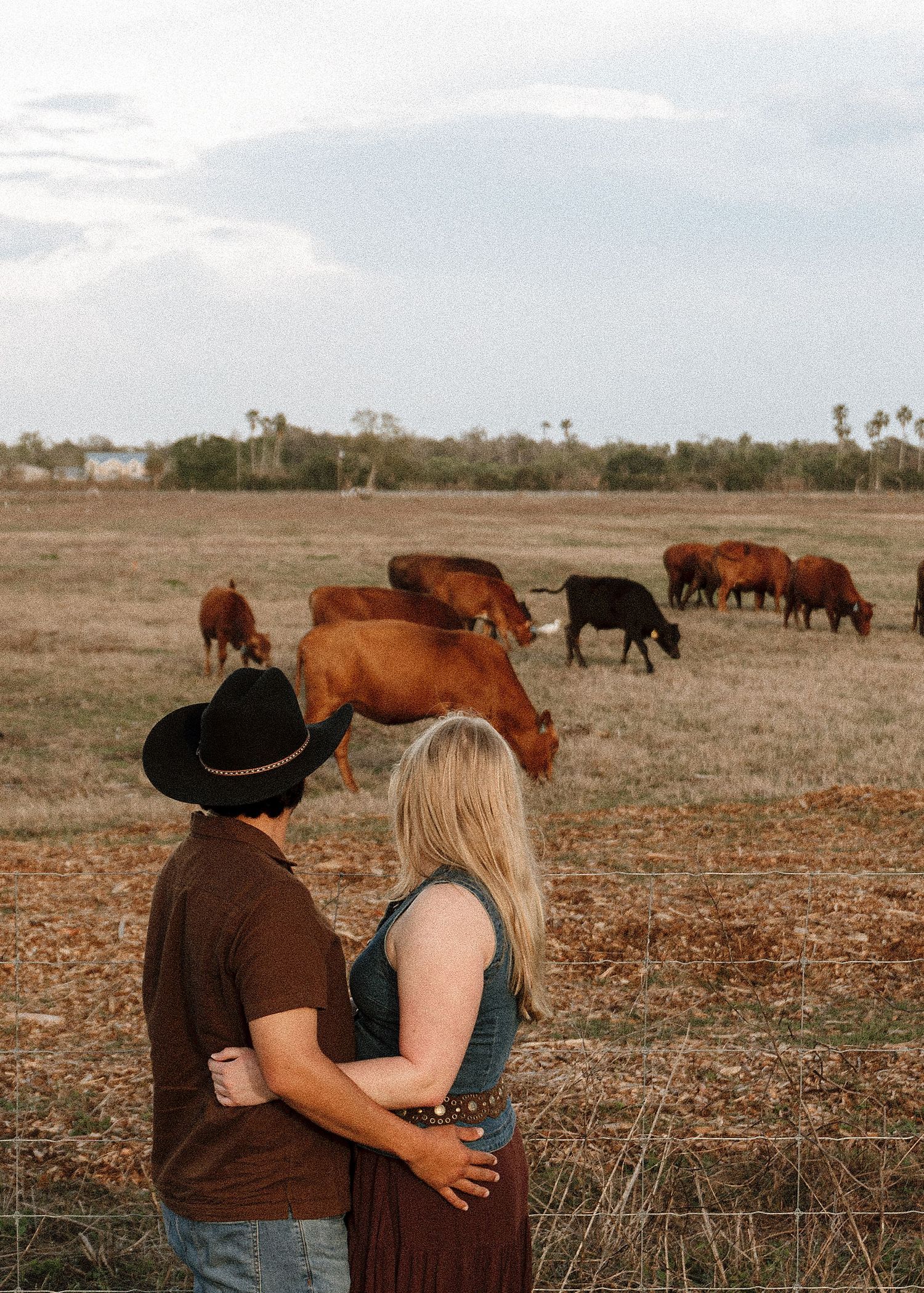 Couple embracing while watching cattle graze on a rustic farm field under a cloudy sky.