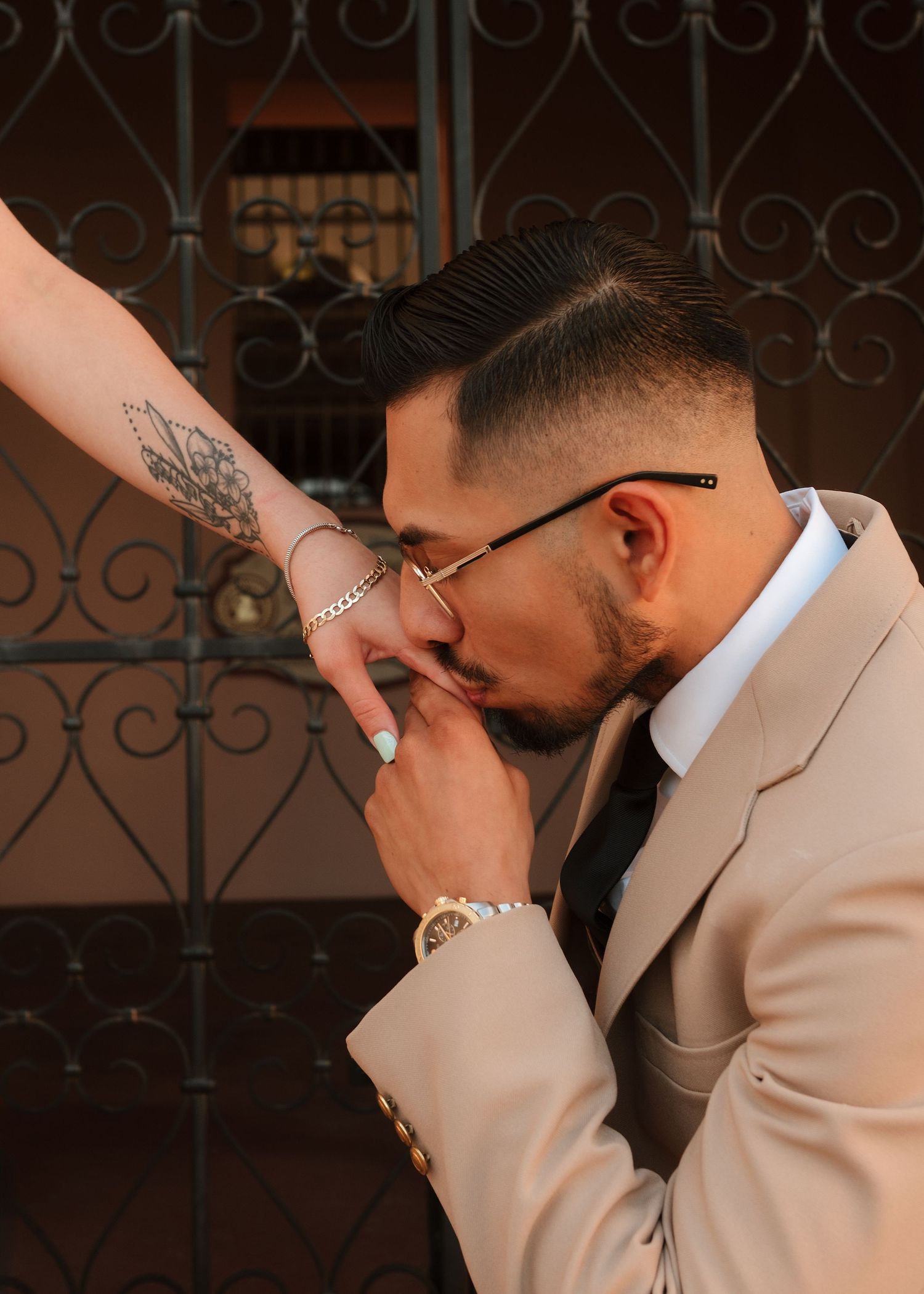 A man in a beige suit kisses a tattooed woman's hand near an ornate iron gate.