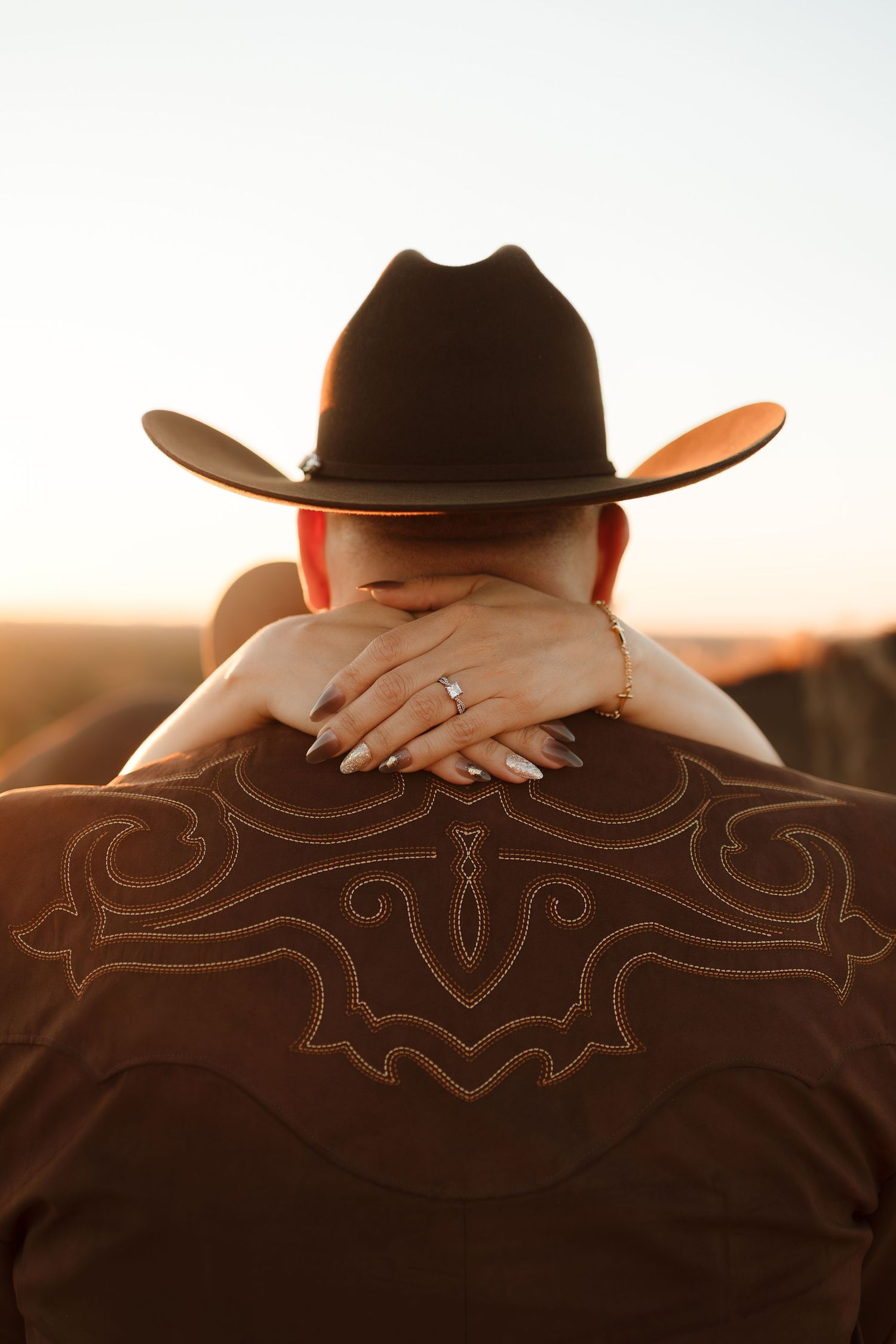 A person in a brown cowboy hat and embroidered western shirt at sunset with a woman's hands clasped around their neck.