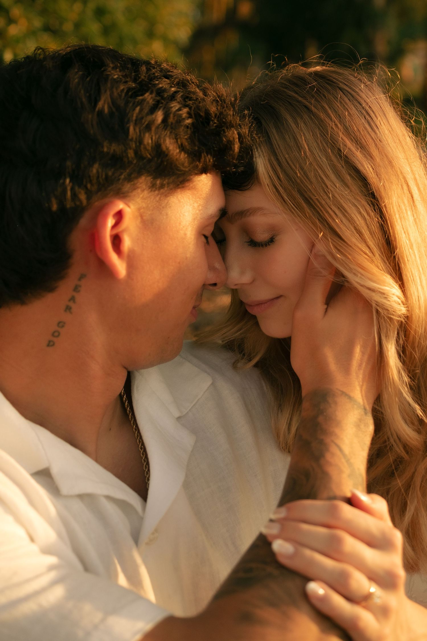 A couple with tattoos share an intimate forehead-to-forehead moment in warm golden sunset light outdoors.