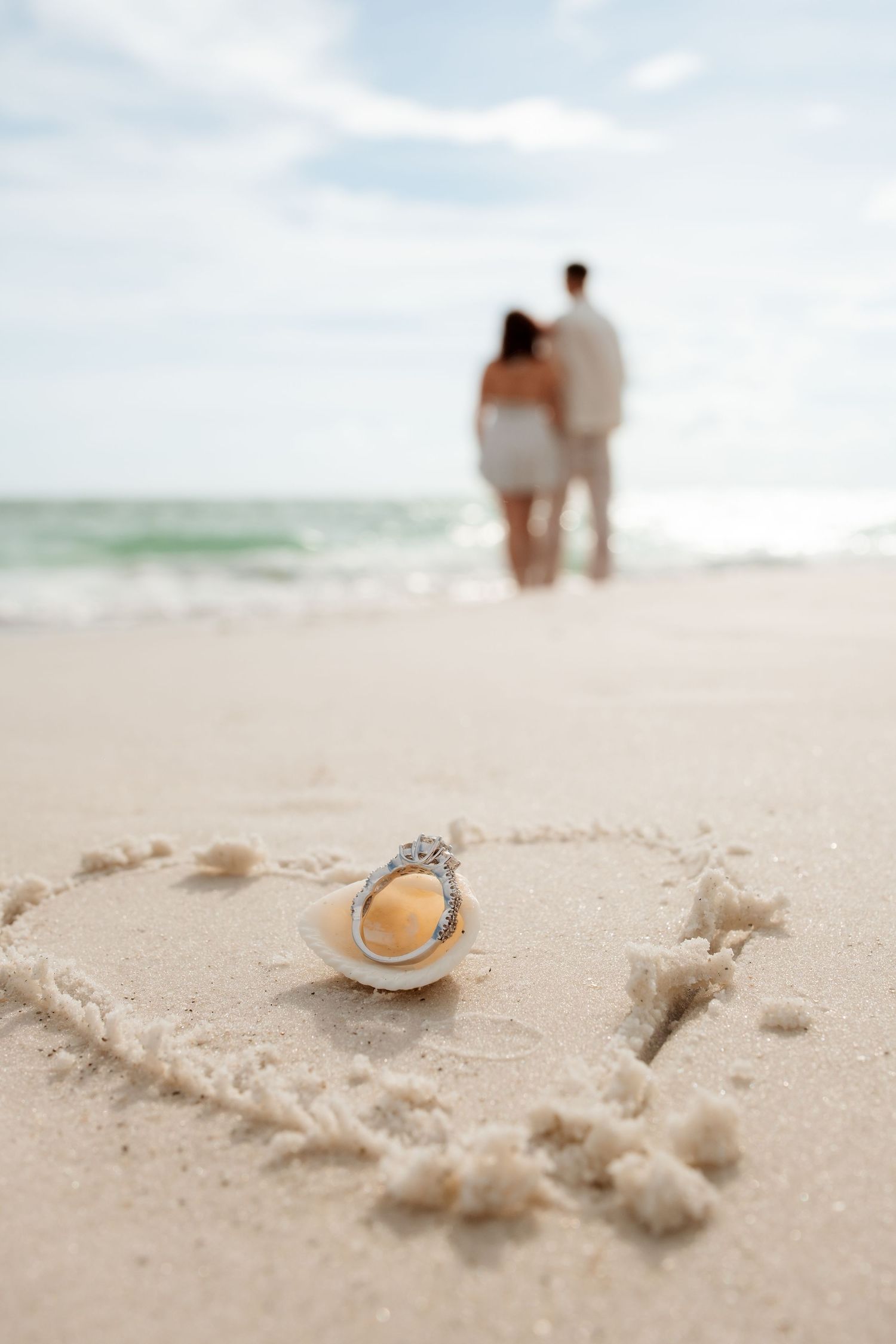 Couple standing at ocean shore with engagement ring in seashell inside heart drawn in sand on beach.