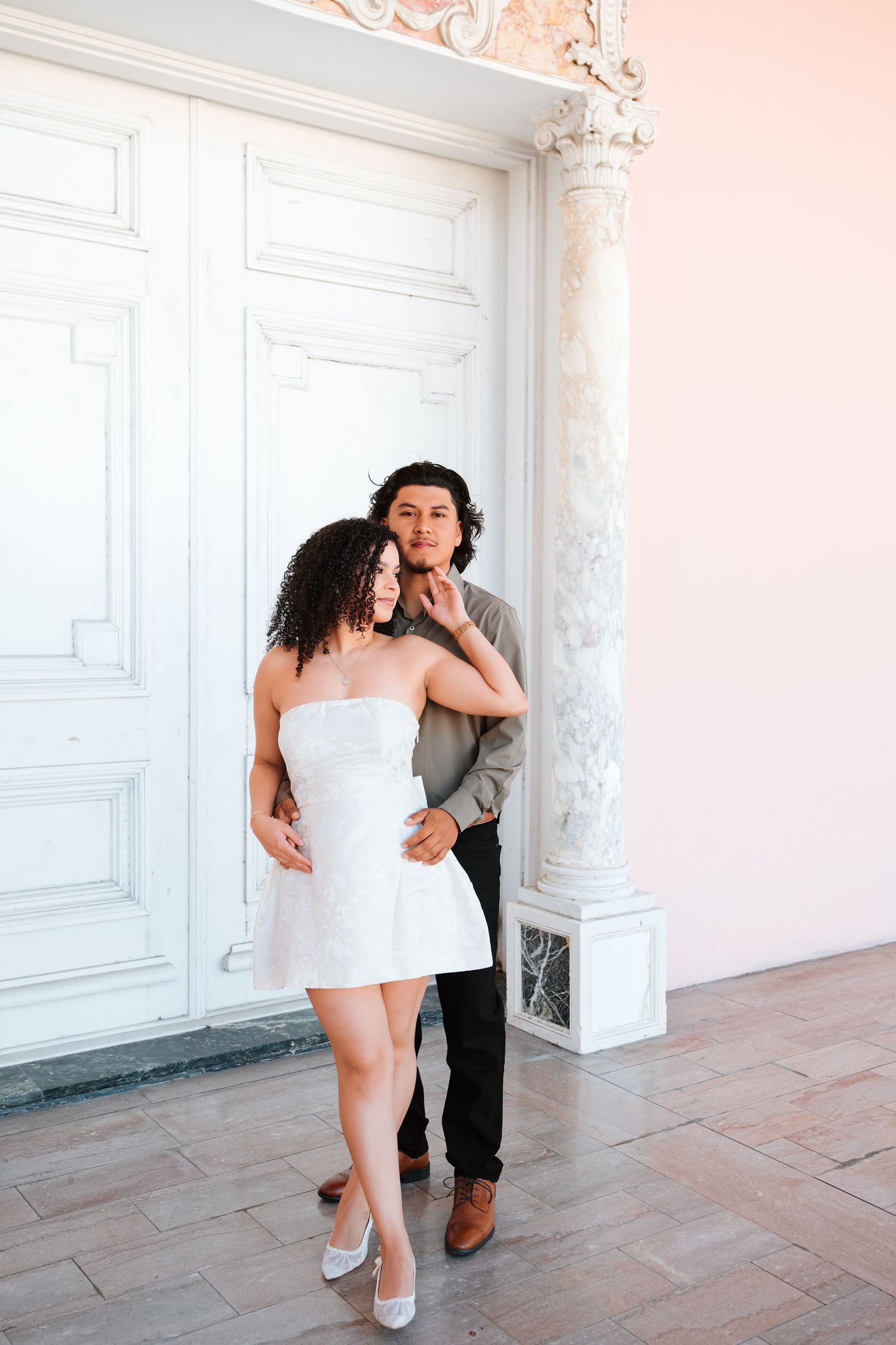 Couple posing together in front of ornate white door with decorative columns, woman in white strapless mini dress.