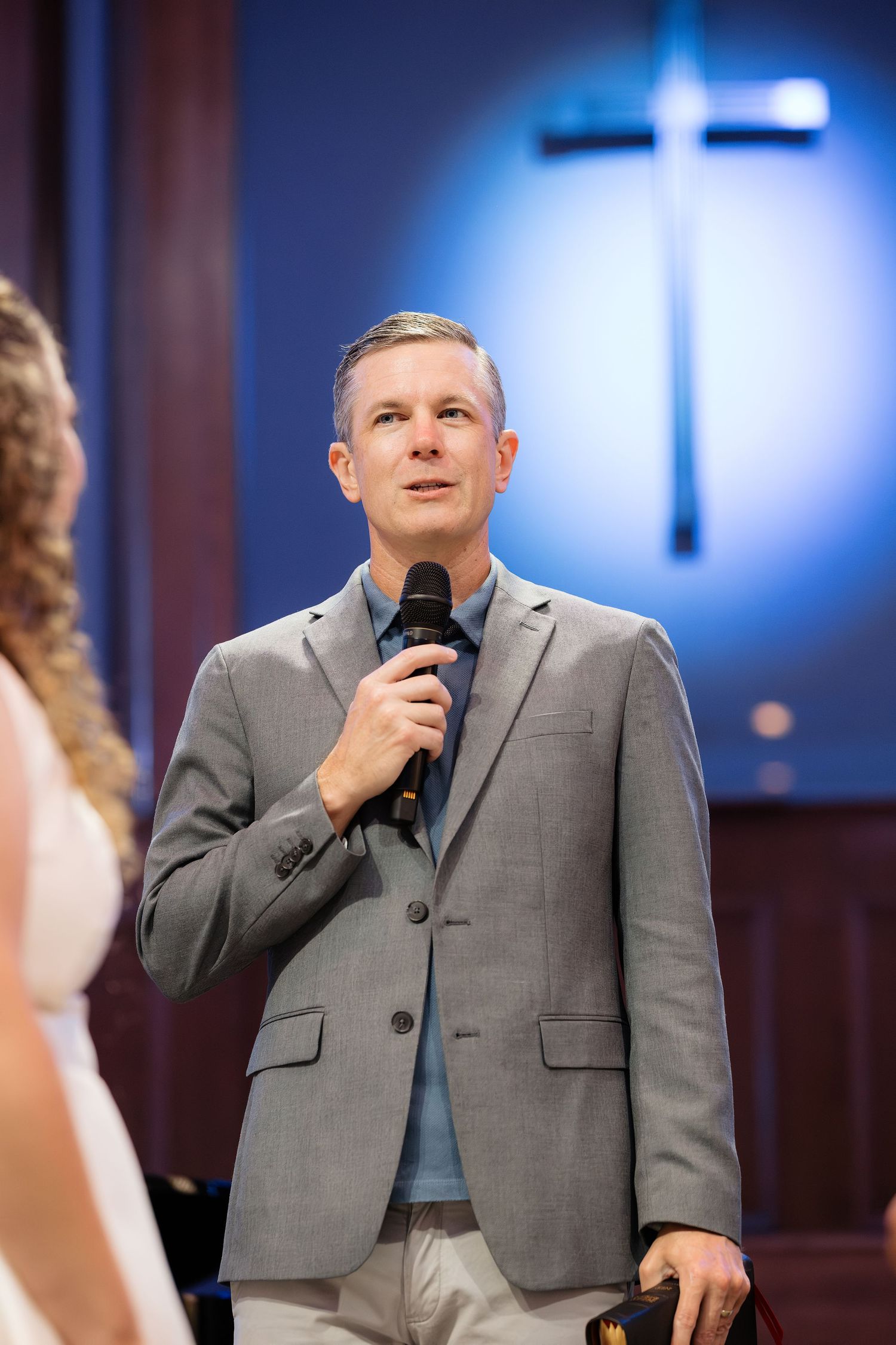 Man in gray suit holding microphone speaks at church podium with illuminated cross in background.