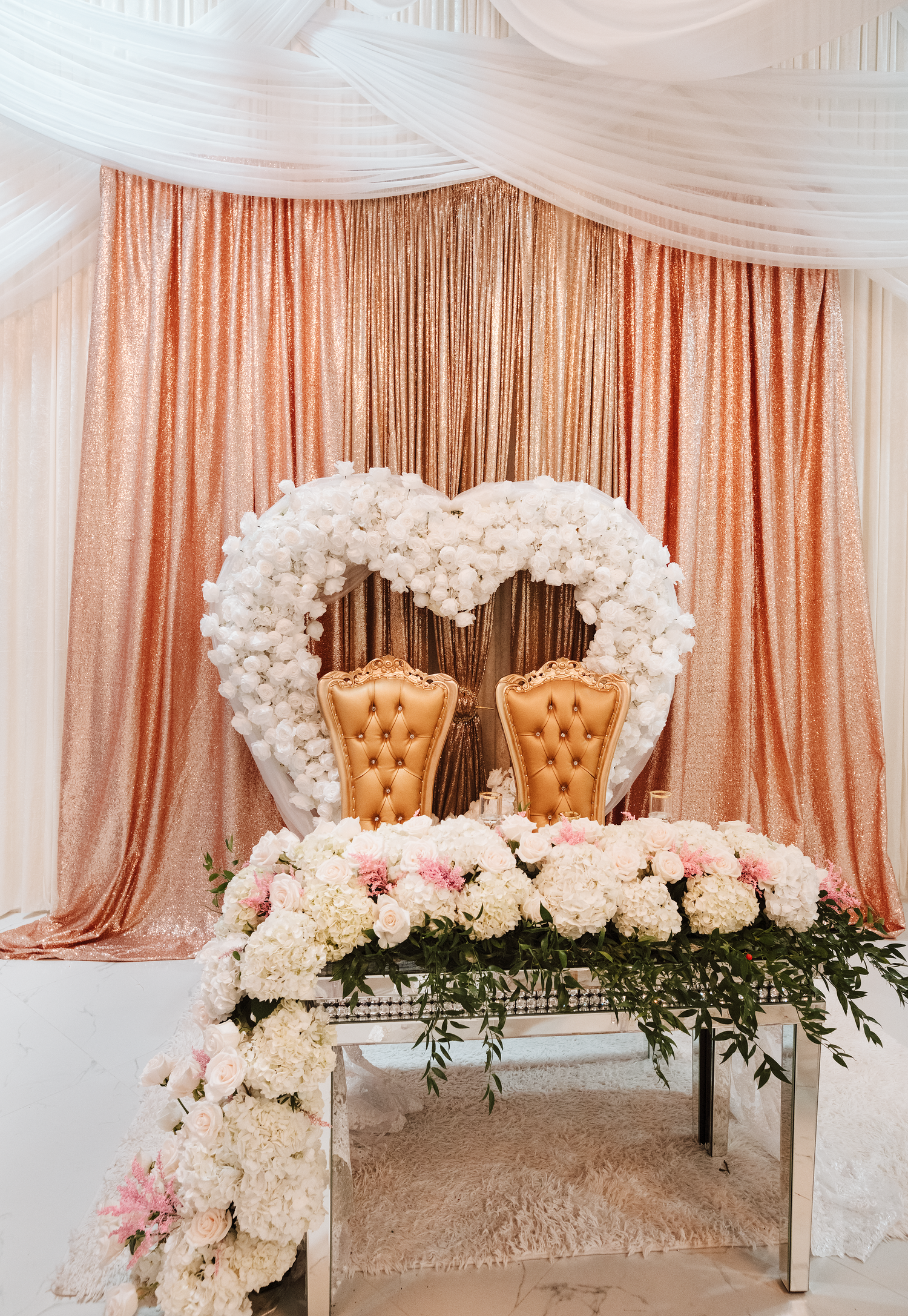 Elegant wedding sweetheart table with gold tufted chairs, heart-shaped floral arch, and white blooms against rose gold sequin backdrop.