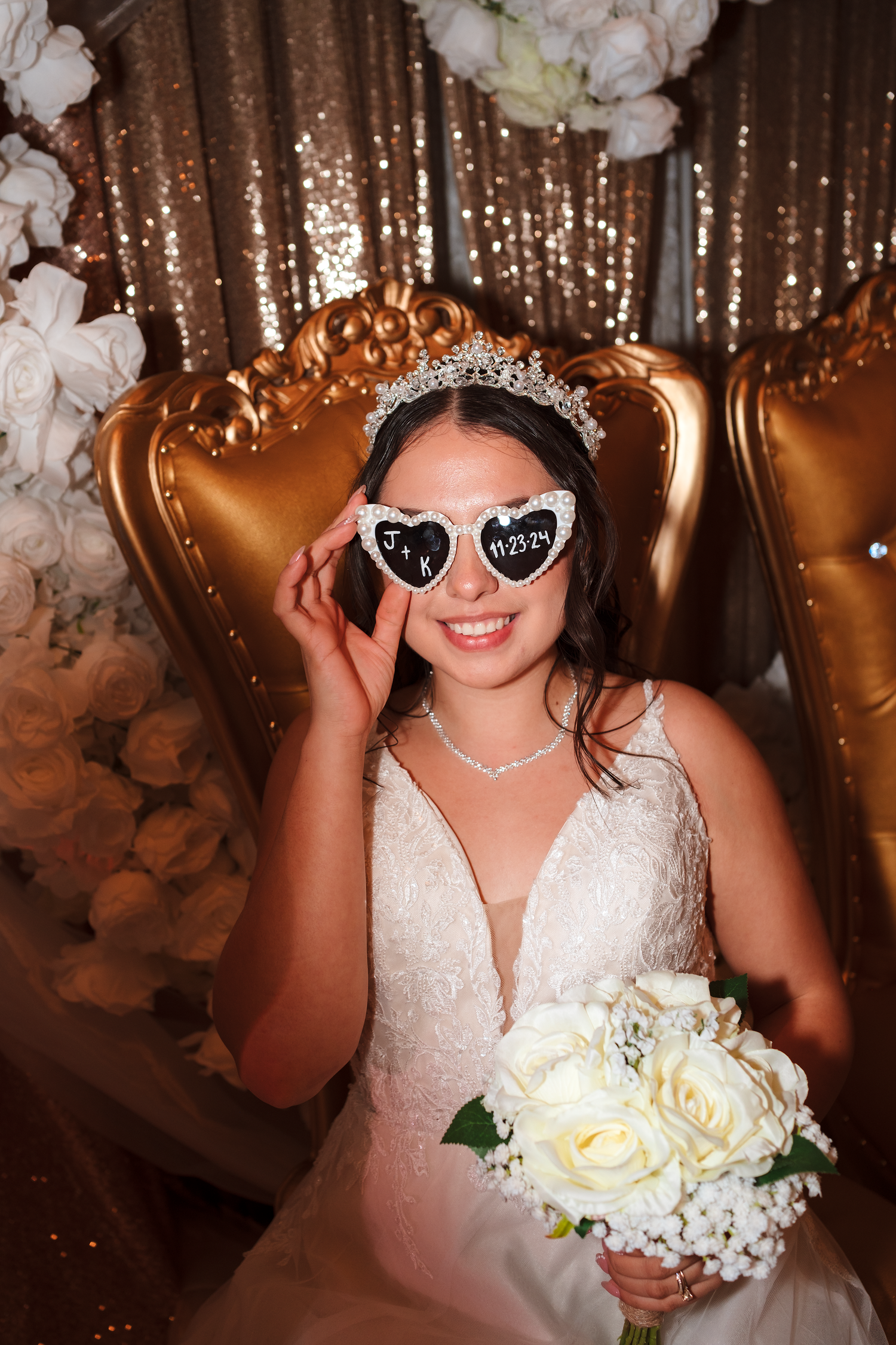 Smiling bride in white lace gown wearing heart-shaped sunglasses and tiara, holding white rose bouquet on gold throne chair.