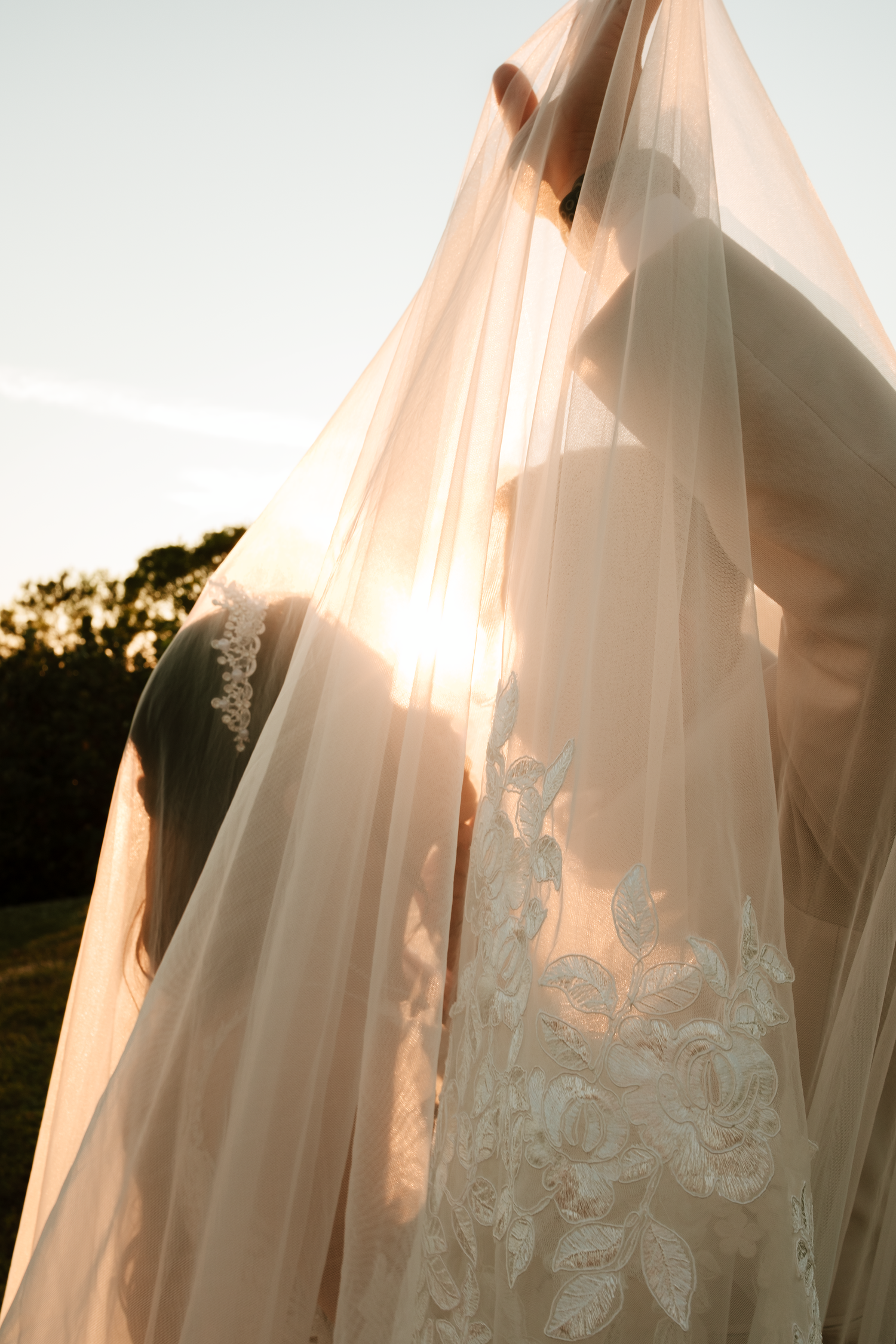 Romantic backlit wedding photo of bride lifting sheer veil against glowing sunset sky with lace gown detail.