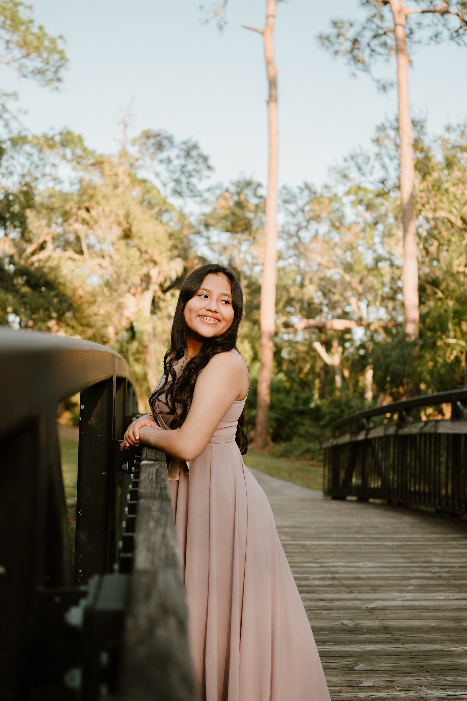 Smiling young woman in a flowing blush pink gown leans on a wooden bridge railing surrounded by tall trees in warm golden light.
