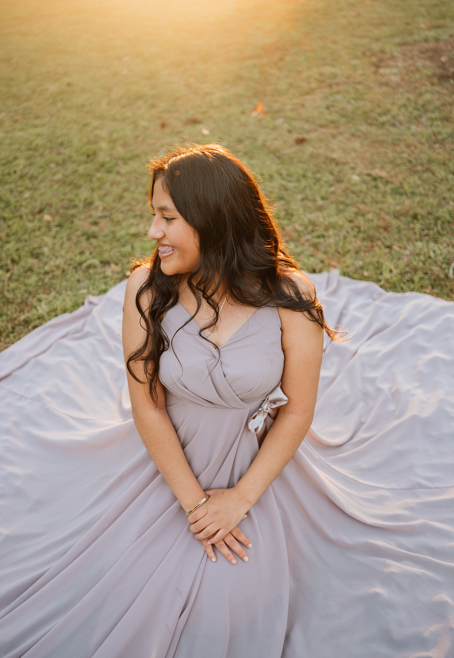 Smiling woman with long wavy dark hair sits on grass in a flowing lavender gown spread around her, glowing in warm golden sunset light.