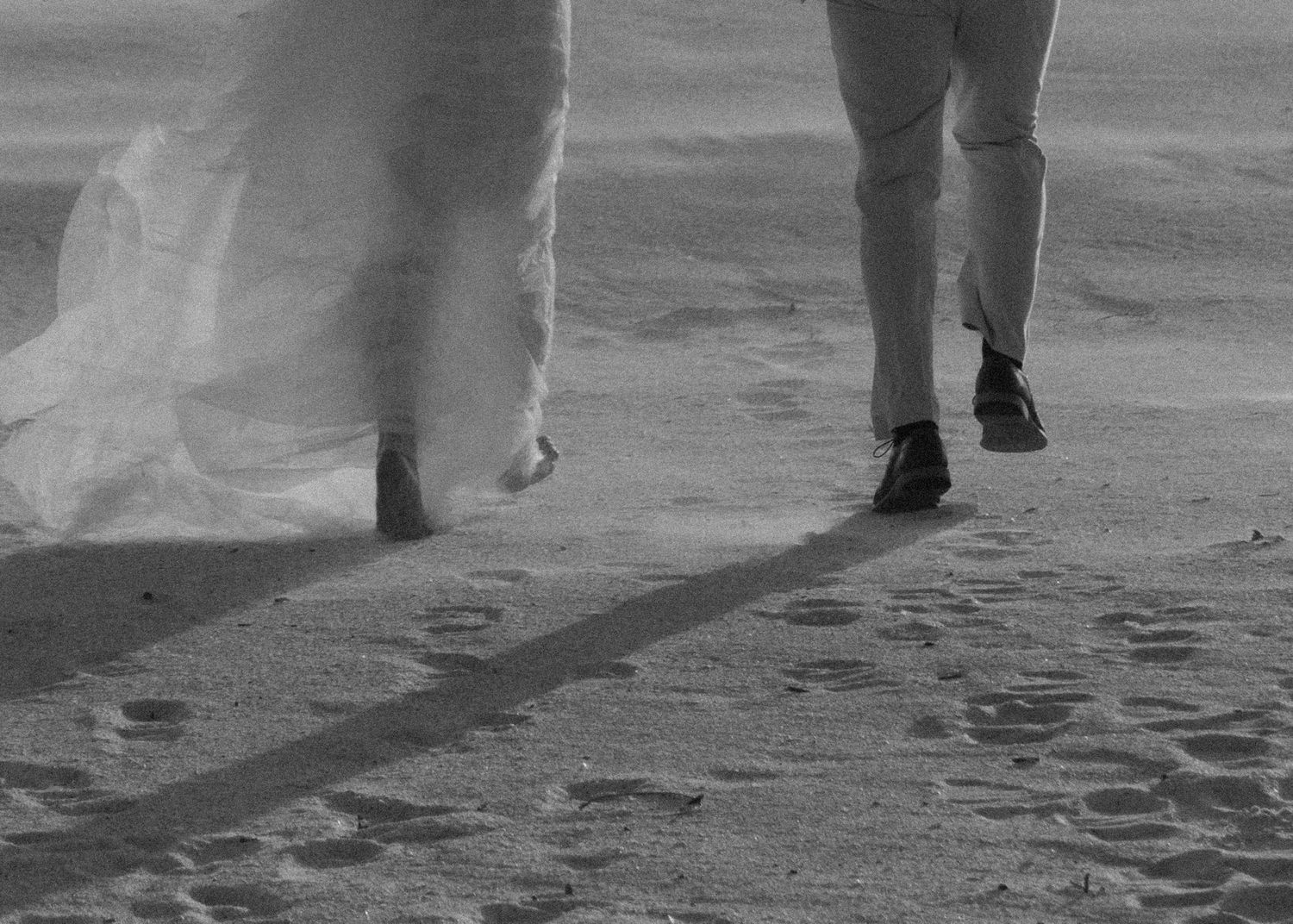 Black and white photo of a bride and groom walking on sandy beach, showing legs and footprints in sand.