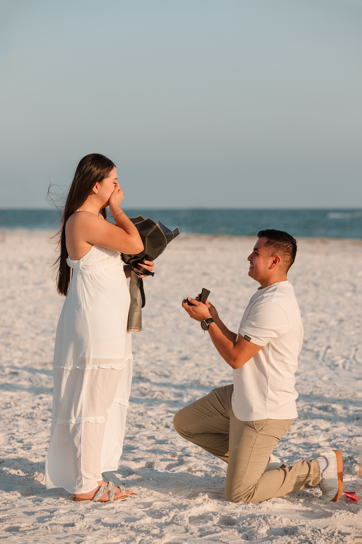 Man proposes on beach at sunset, kneeling with ring box as woman in white dress covers her face in surprise.