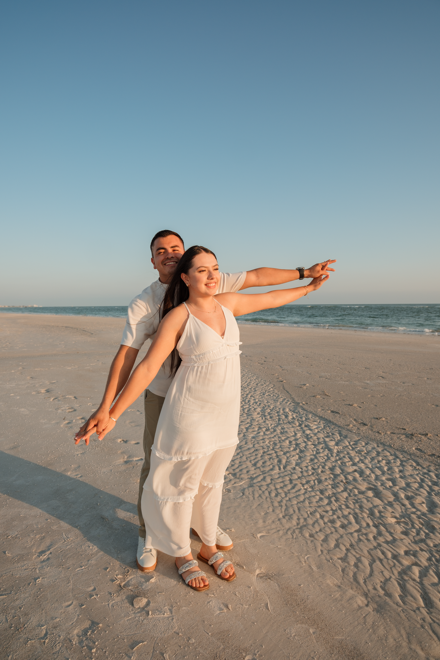 Happy couple posing on a sandy beach at sunset, woman in white dress pointing toward the ocean horizon.