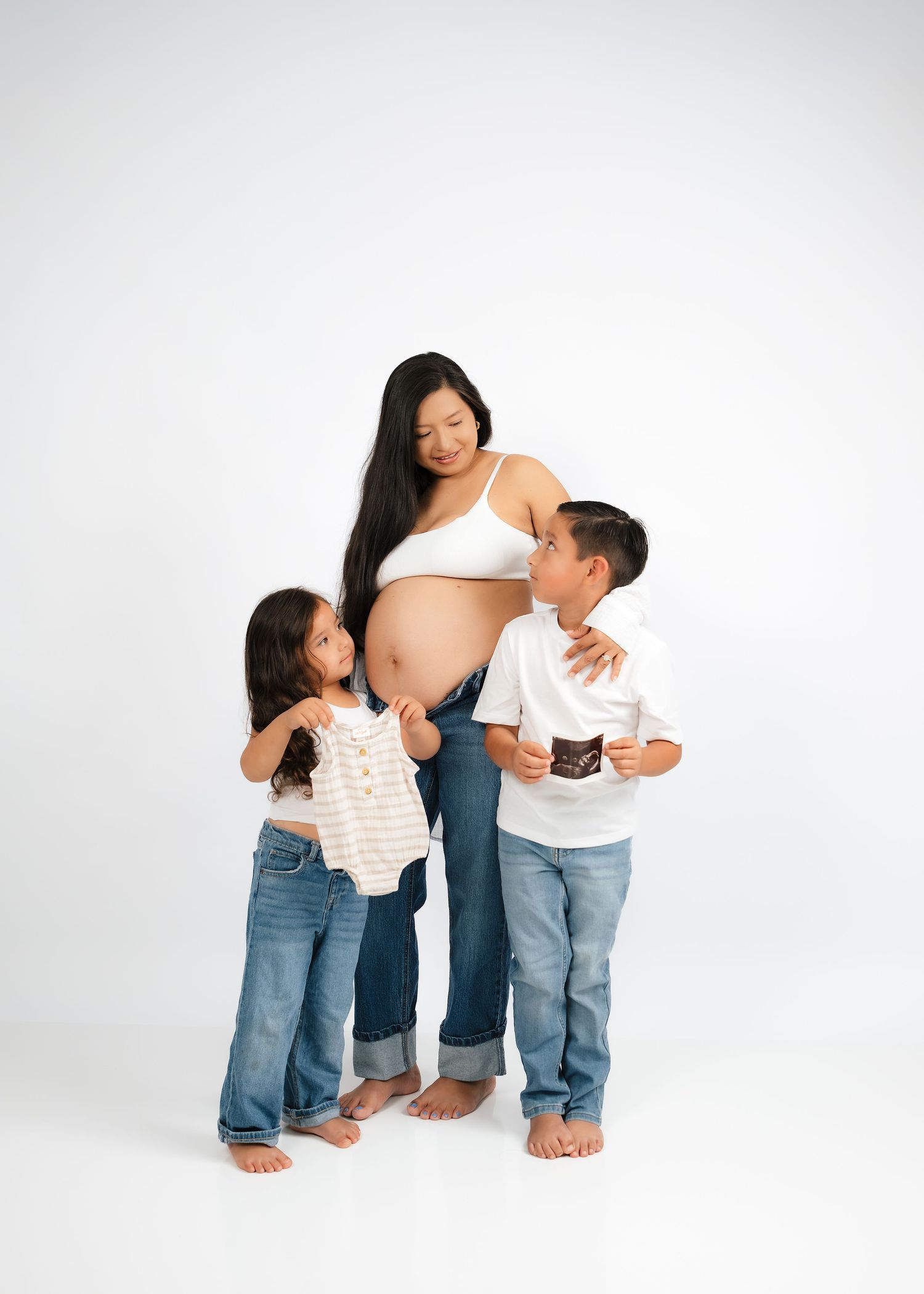 Pregnant woman in white crop top and jeans poses with two young children holding baby clothes and sonogram photo.