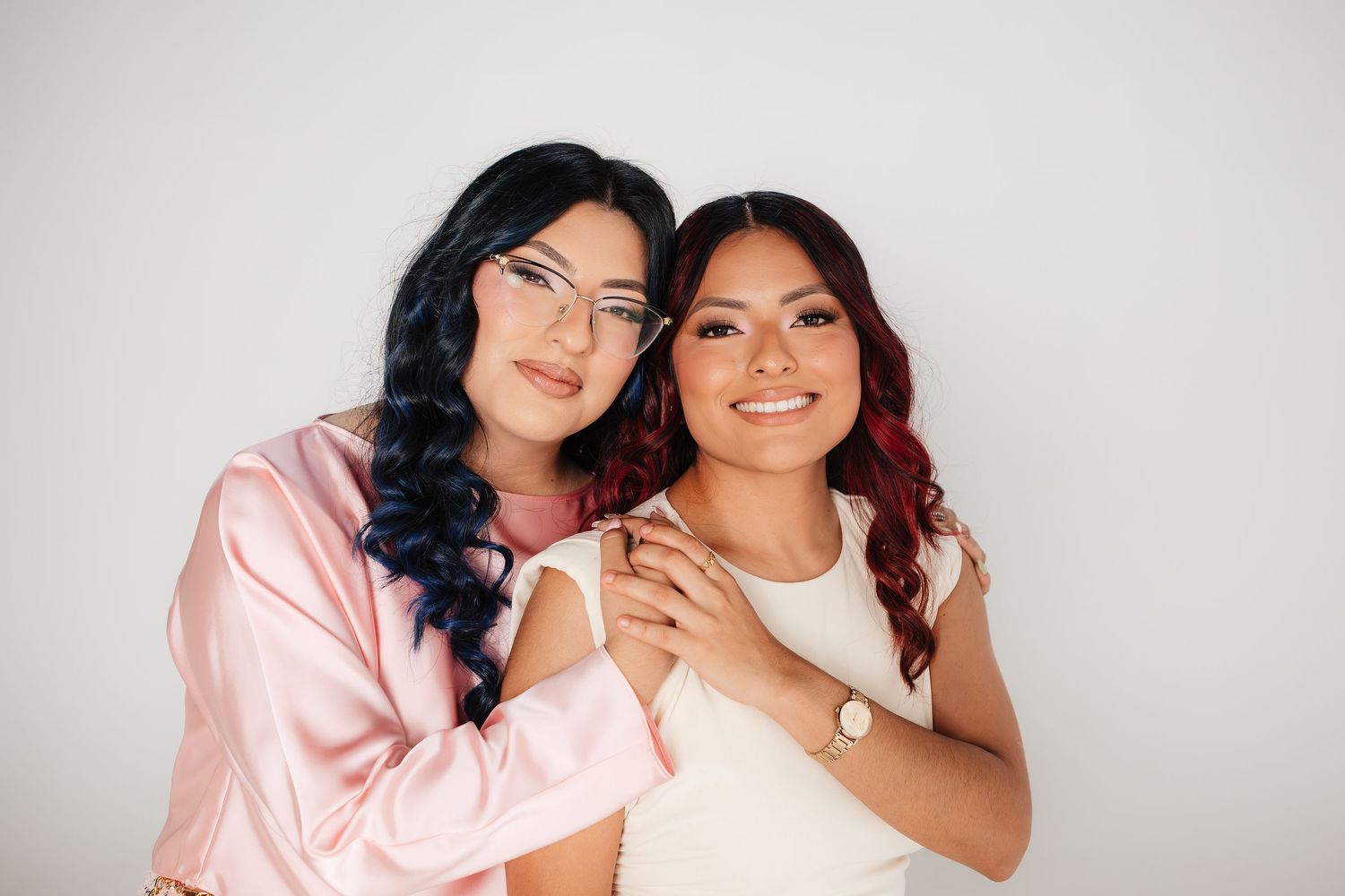 Two smiling women pose together, one in pink satin and glasses, the other in white, against a light background.