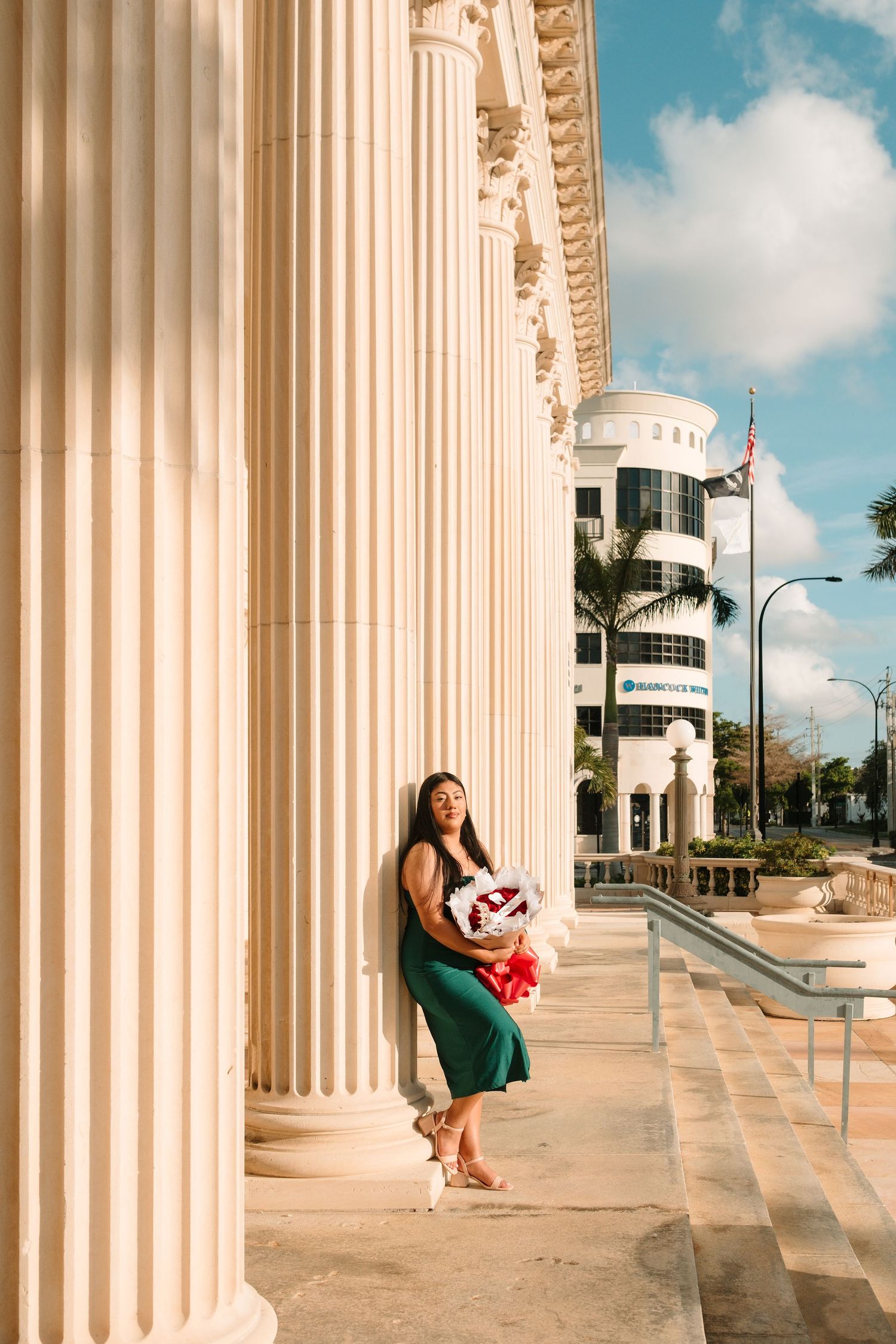 Woman in green skirt holding flowers leans against white columns of a classical building on a sunny day.