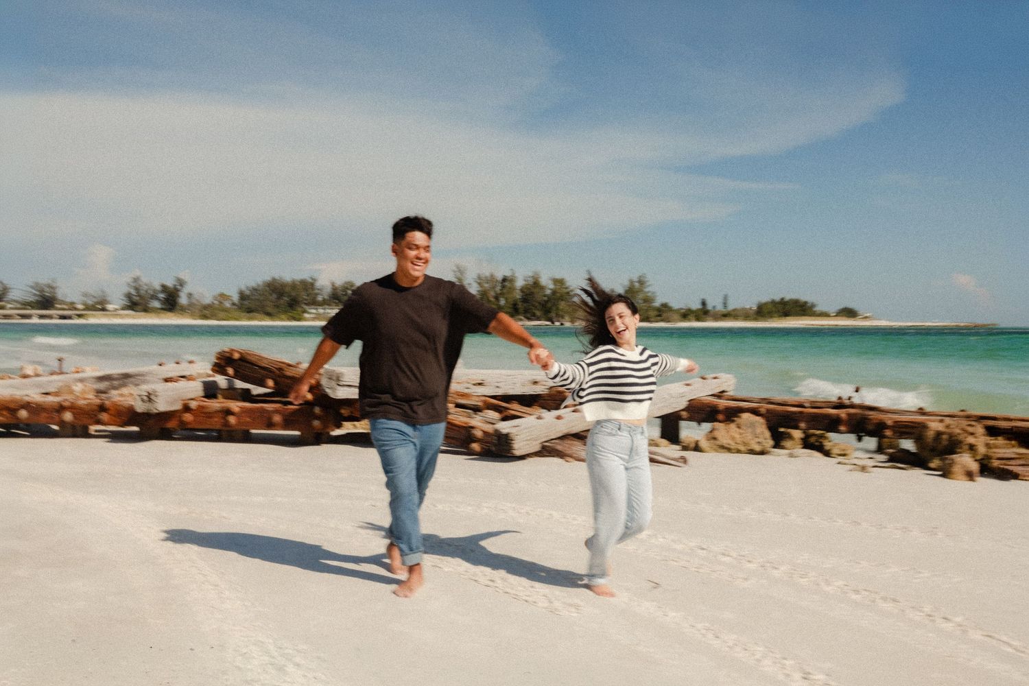 A happy couple holds hands and runs barefoot on a white sandy beach with turquoise water and blue skies.