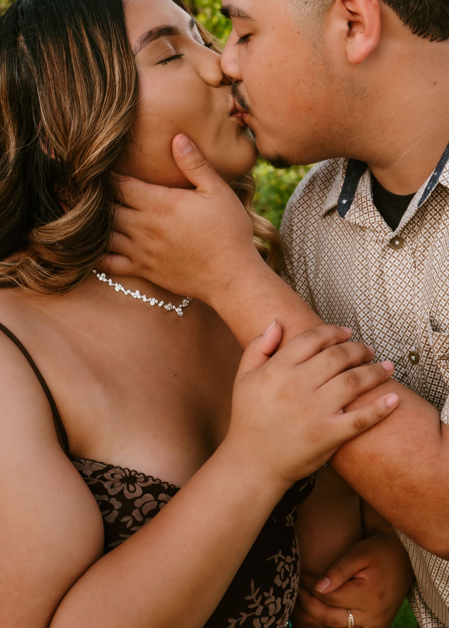 A couple shares a romantic kiss, the woman wearing a delicate necklace while holding hands tenderly outdoors.