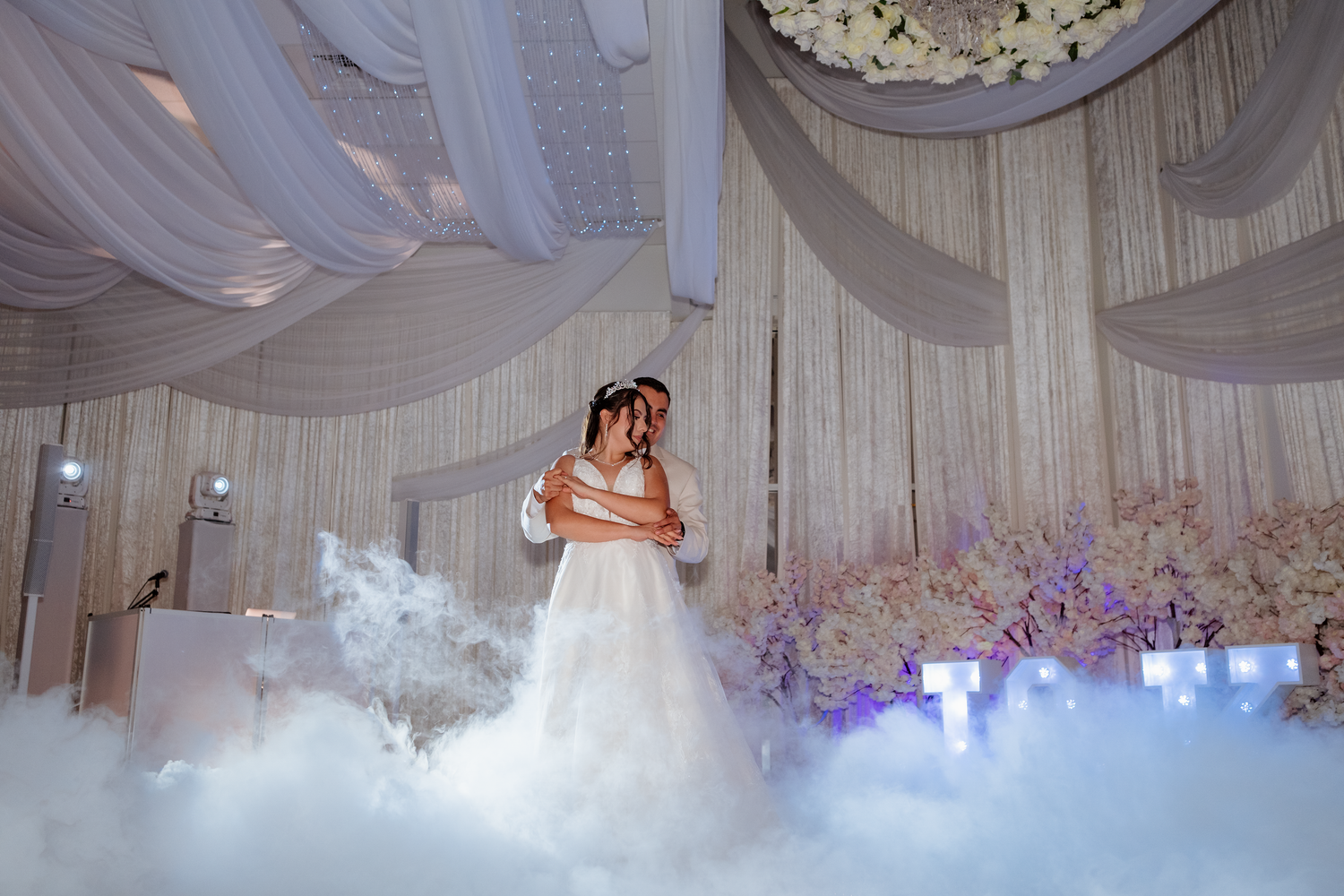 Bride and groom share first dance surrounded by dramatic fog on a white floral wedding reception stage.